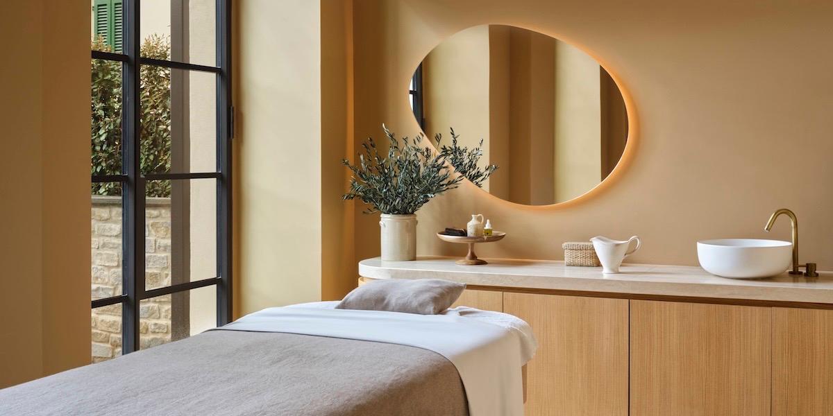 a spa treatment bed with folded linens beneath a round mirror, a wooden cabinet holding a vase of olive branches, and a door open to a stone courtyard