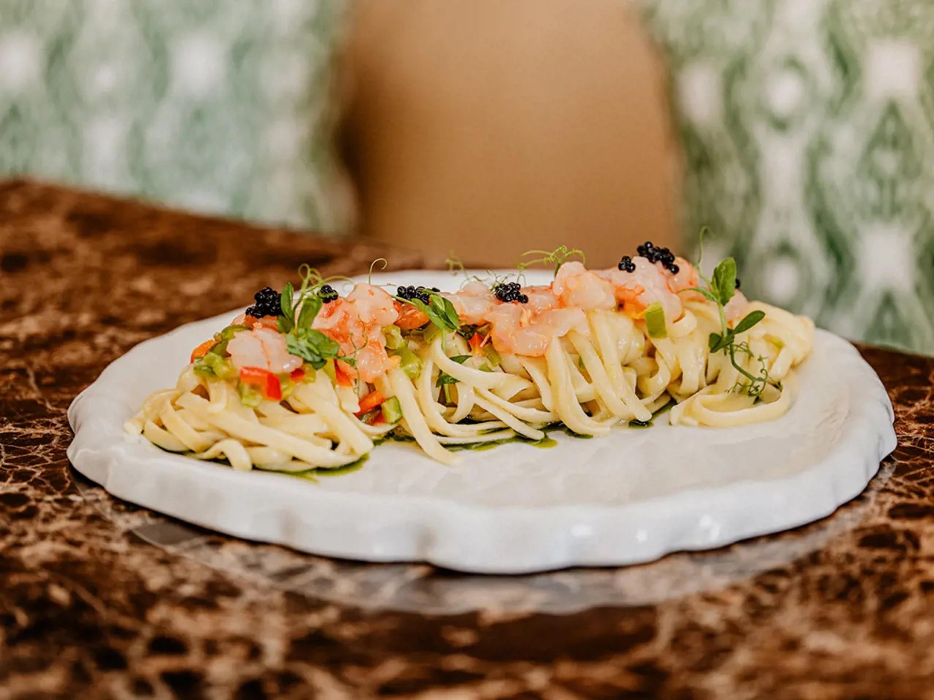 A plate of linguine with shrimp, caviar and herbs on a white ceramic plate, set on a marble table with a blurred figure seated behind.