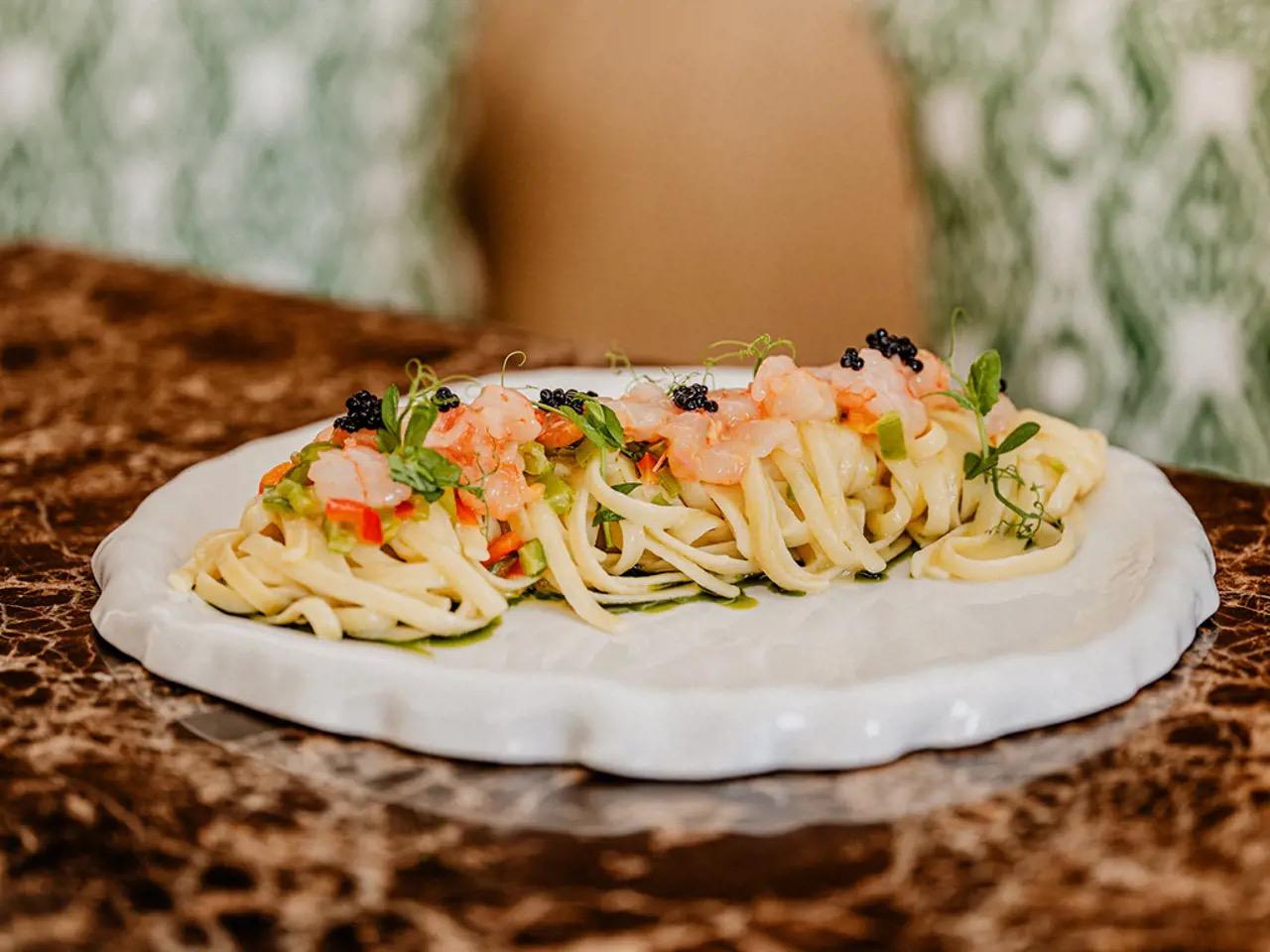 A plate of linguine with shrimp, caviar and herbs on a white ceramic plate, set on a marble table with a blurred figure seated behind.