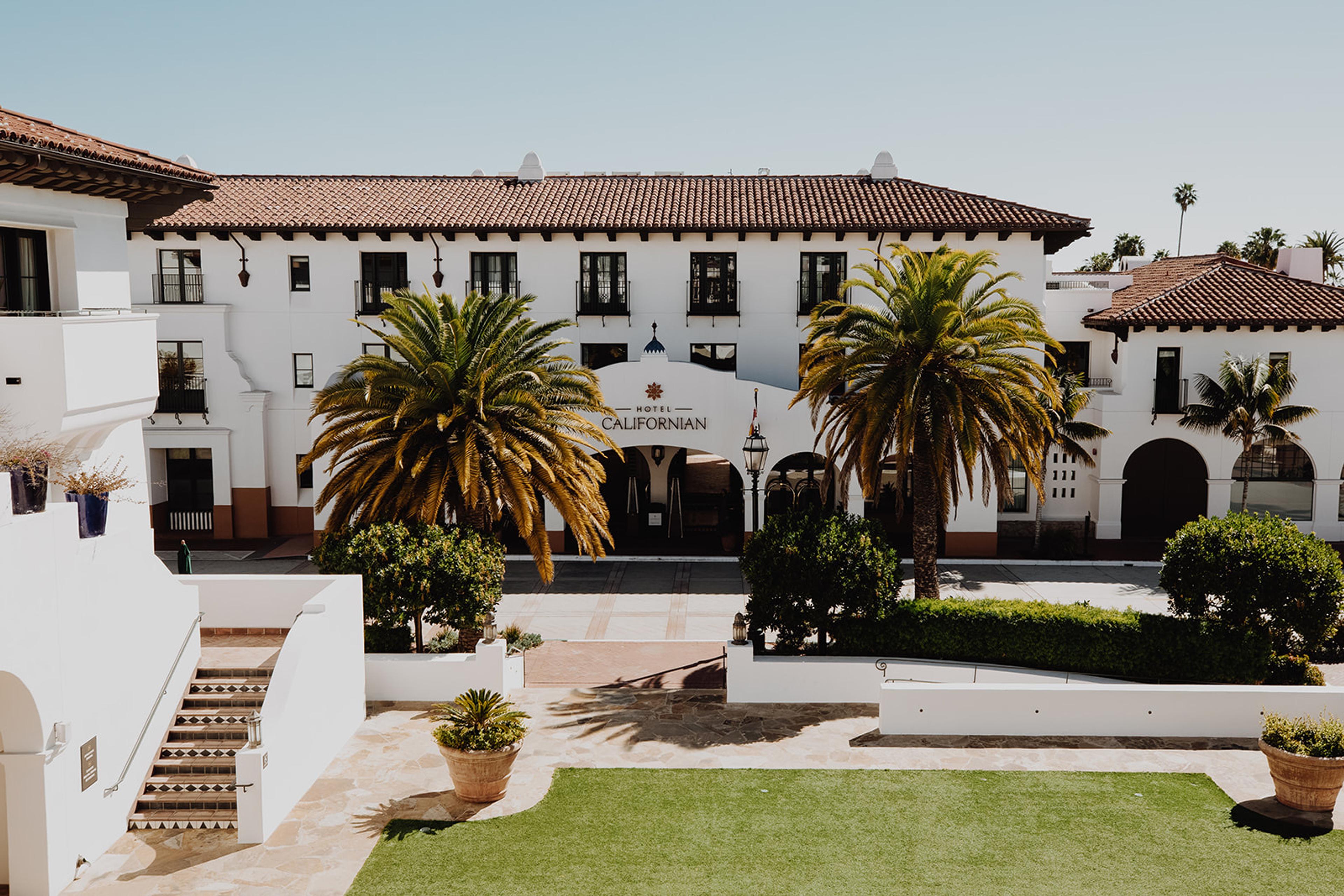 A white building with a red tile roof and arched entrance reading Hotel Californian, seen past two palm trees and a lawn from a raised terrace with stone steps.