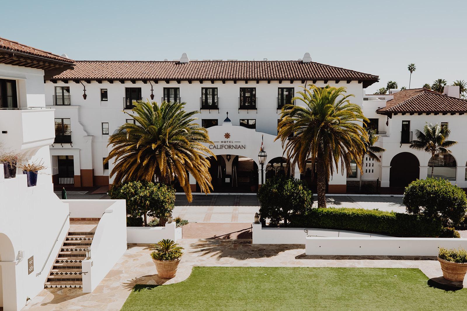 A white building with a red tile roof and arched entrance reading Hotel Californian, seen past two palm trees and a lawn from a raised terrace with stone steps.