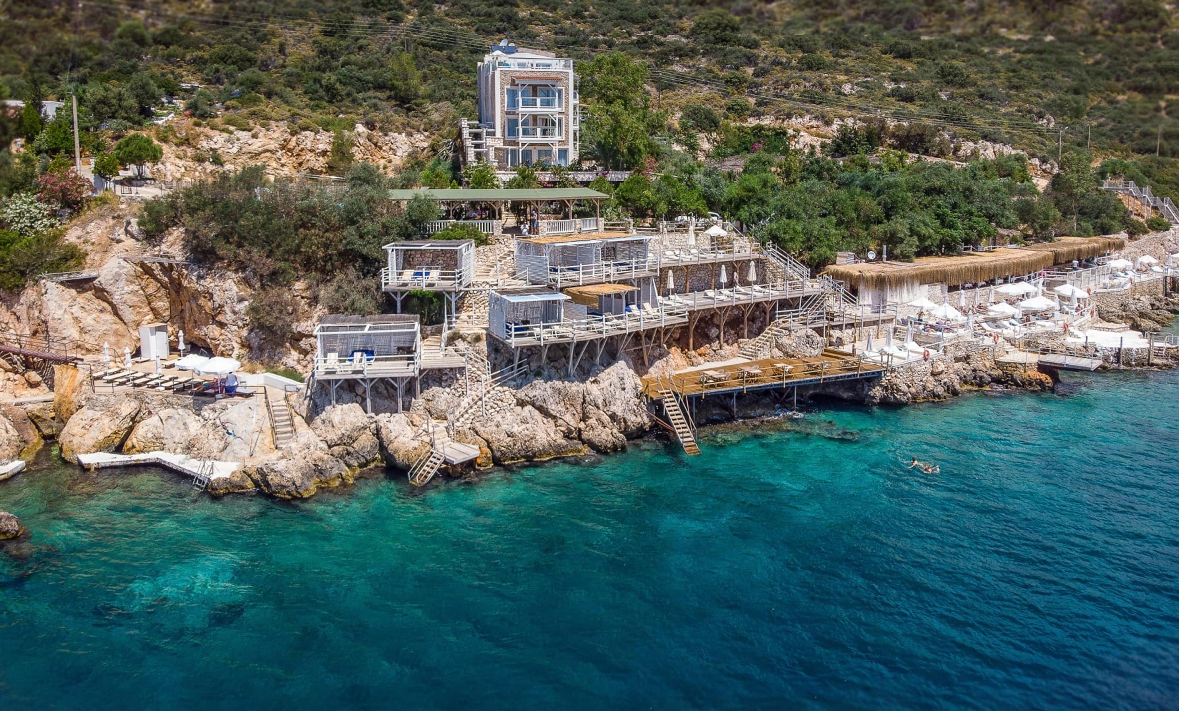 An aerial view of a stone hotel building above a rocky cove, with wooden sundeck platforms stepping down to turquoise water and one swimmer in the distance.