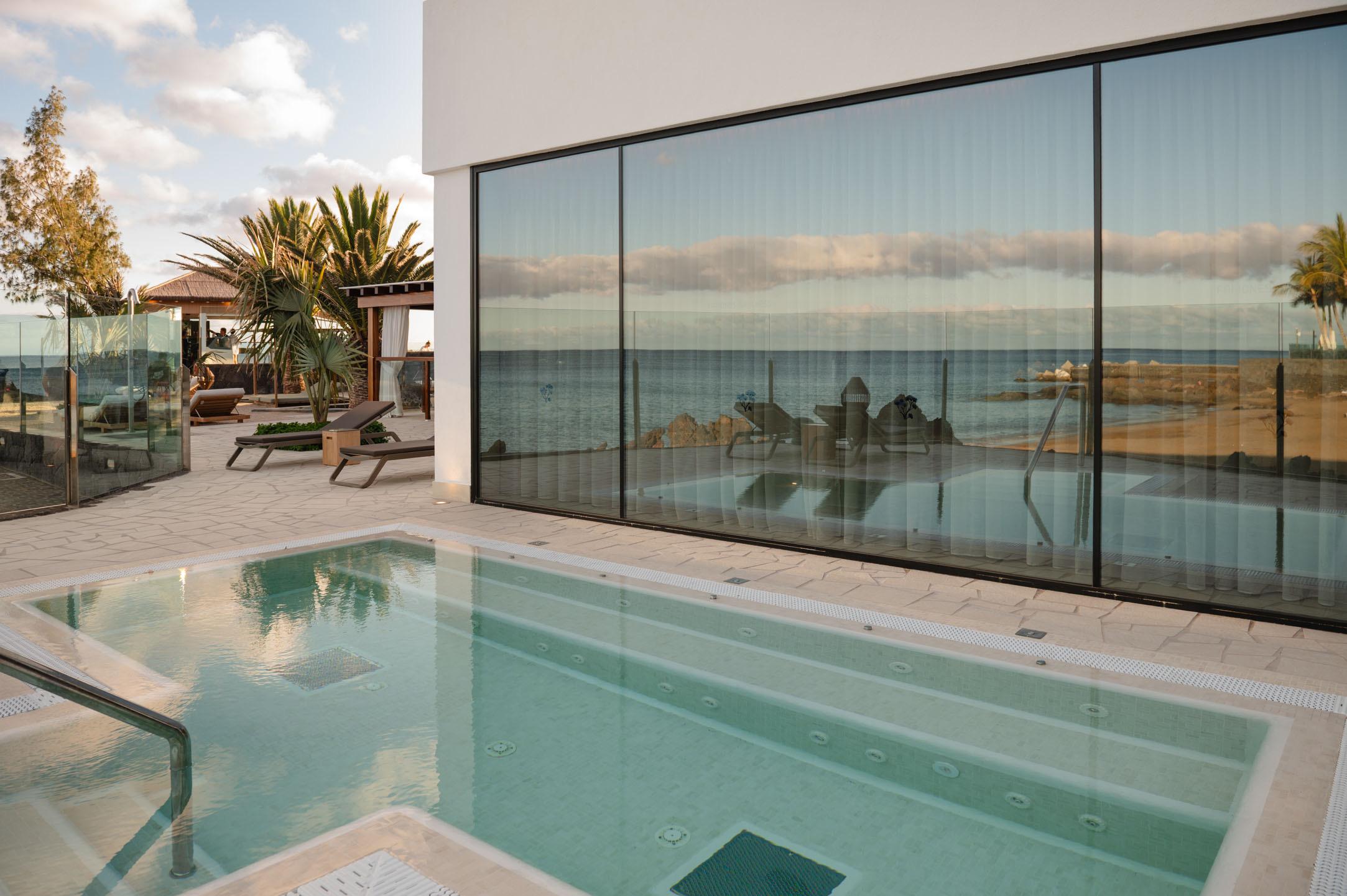 An outdoor spa pool beside a glass-fronted building reflecting the sky, a pergola and loungers on the far side and the sea beyond a low wall.