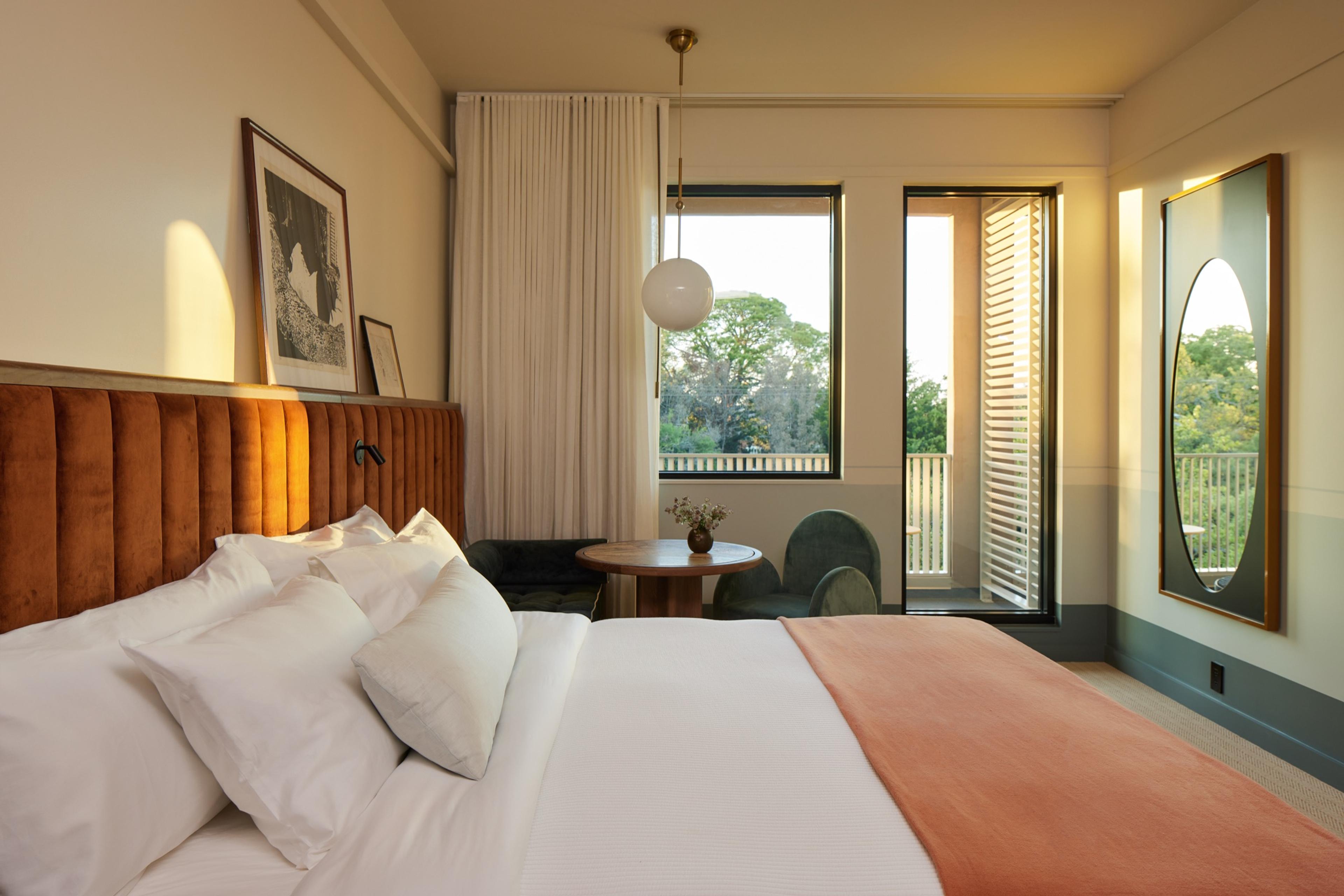 A bedroom with a bed under a rust velvet headboard, a window onto trees and an open balcony door, a round wooden table and a green armchair.