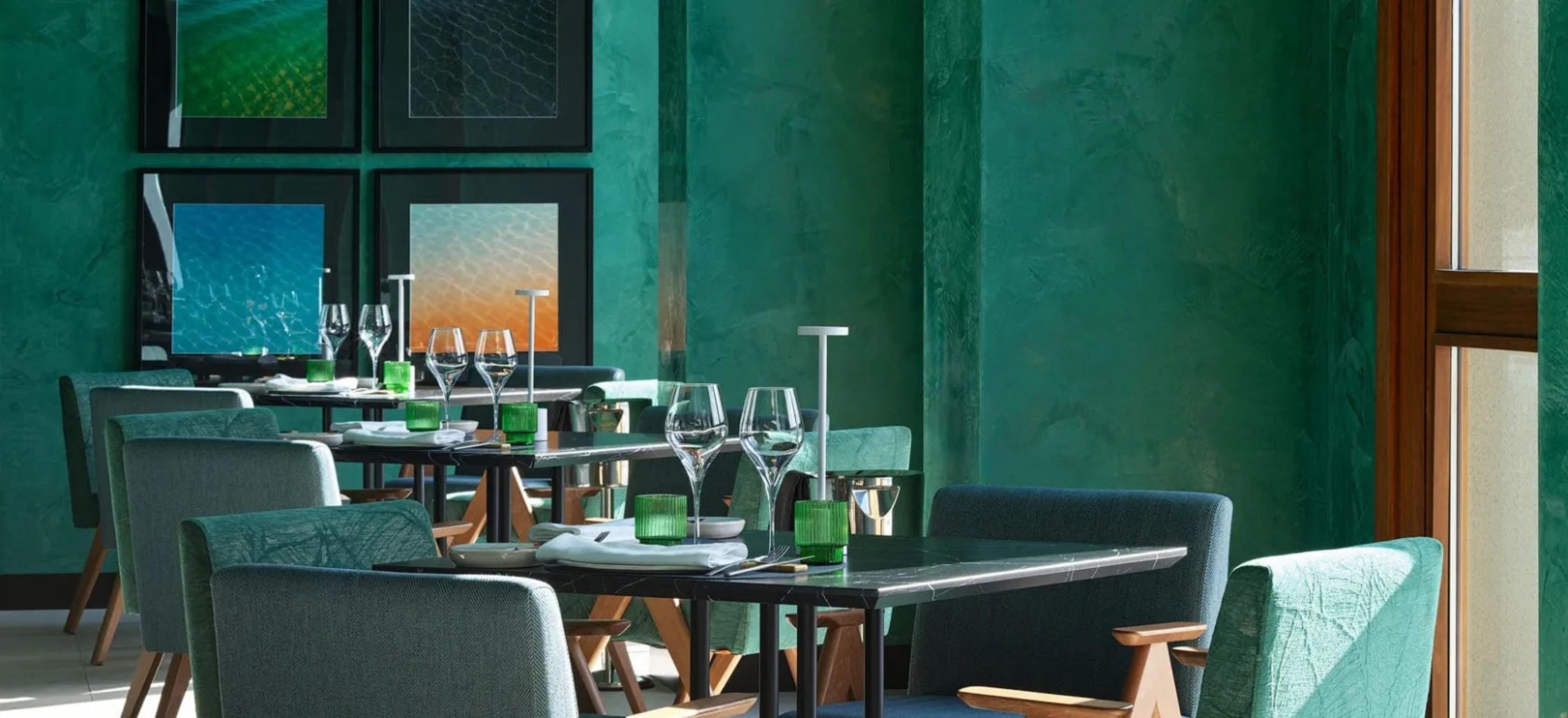 A restaurant interior with teal walls, framed photographs, and tables laid with wine glasses and folded napkins.