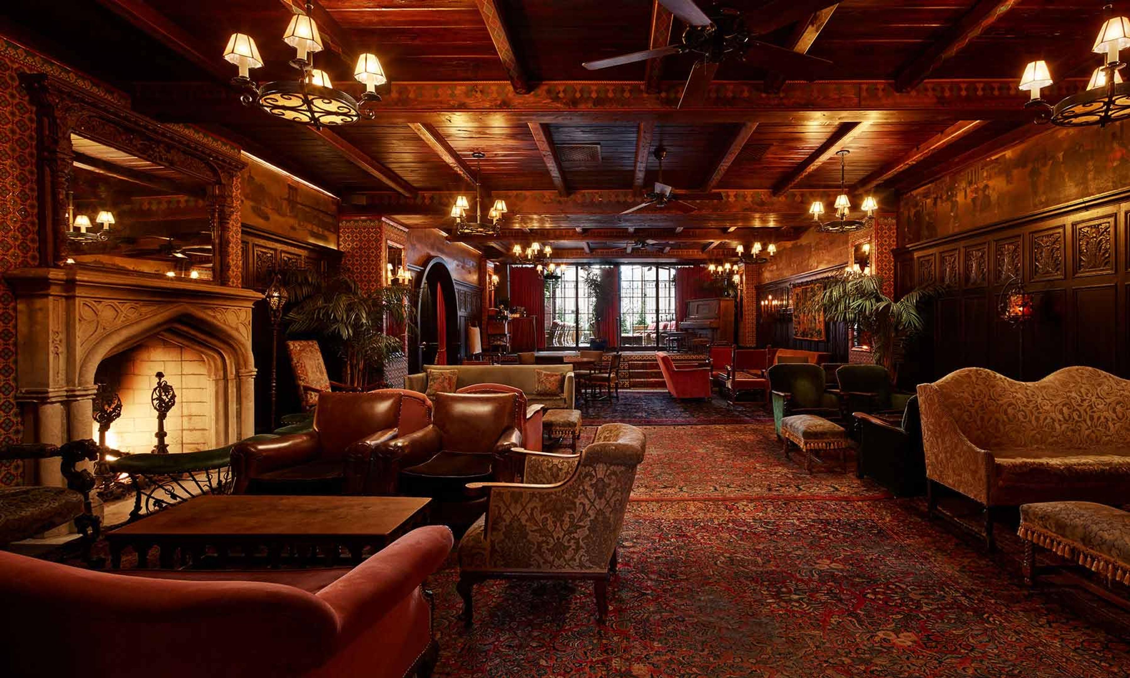 A wood-beamed hotel lounge with leather armchairs, a stone fireplace, patterned rugs and wrought iron chandeliers.