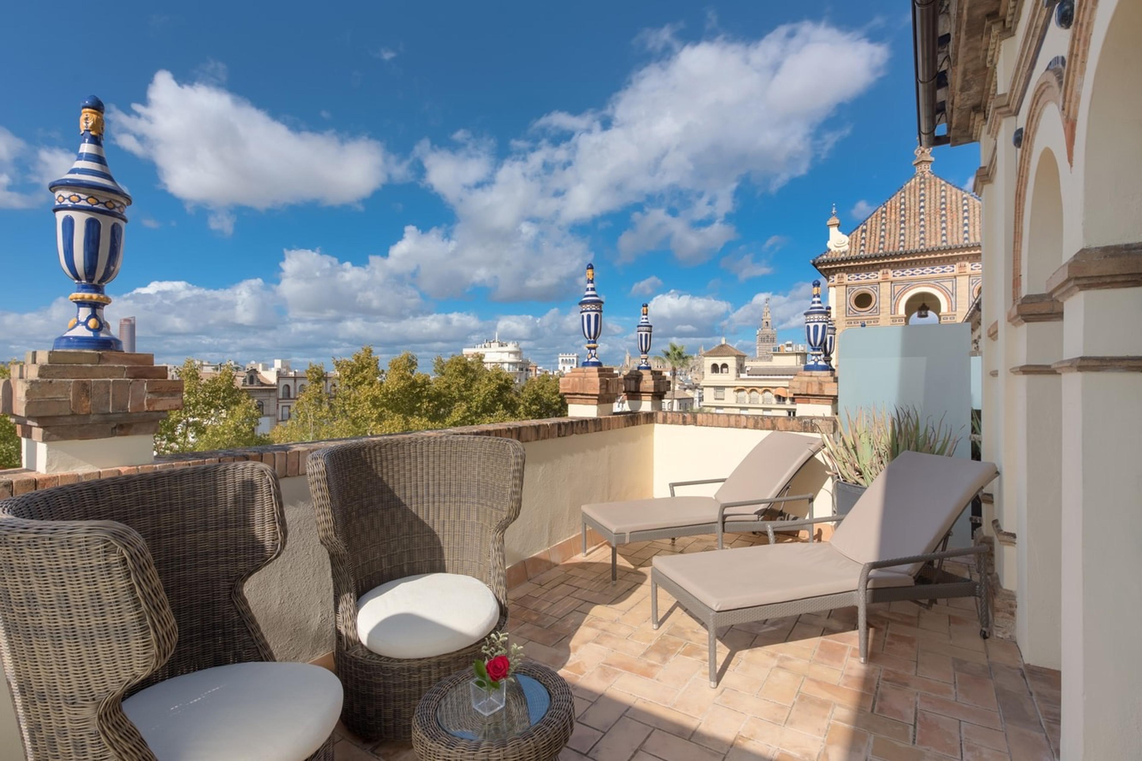 A rooftop terrace with wicker lounge chairs and sun loungers, ceramic finials on the parapet and a tiled tower against a blue sky with clouds.