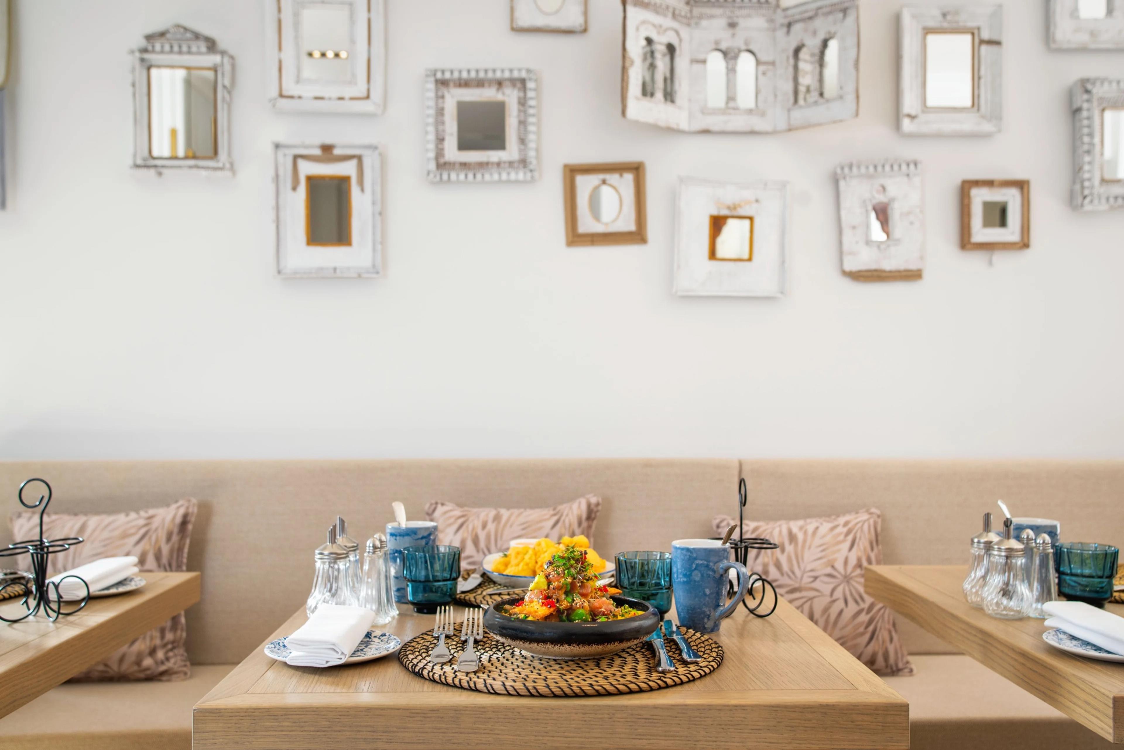 A banquette table set with breakfast dishes, coffee, and fruit, below a wall hung with an assortment of small decorative mirrors.
