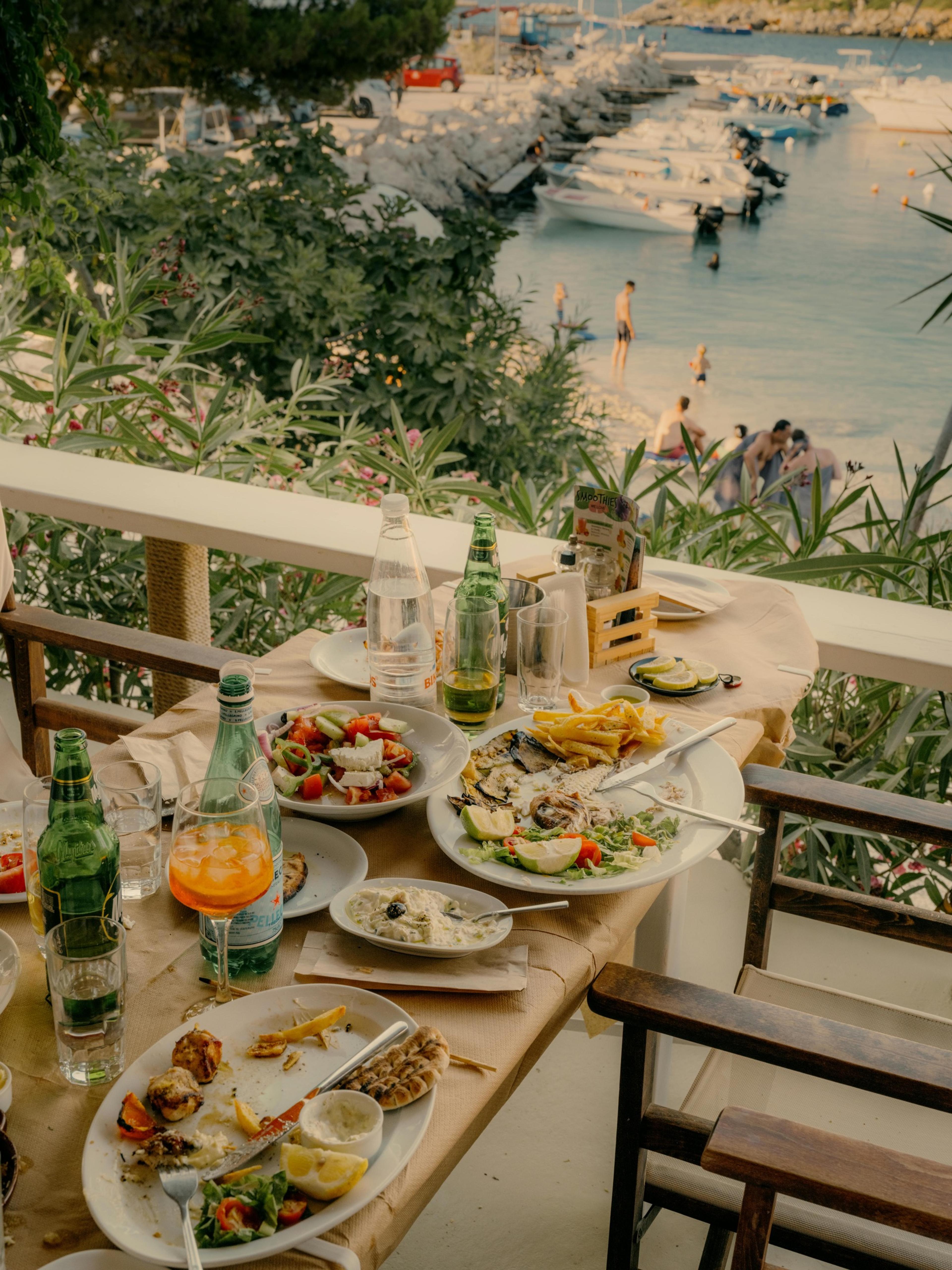 A table set with Greek salad, grilled fish, chips and drinks on a terrace overlooking a marina of moored boats, with swimmers in the sea beyond.