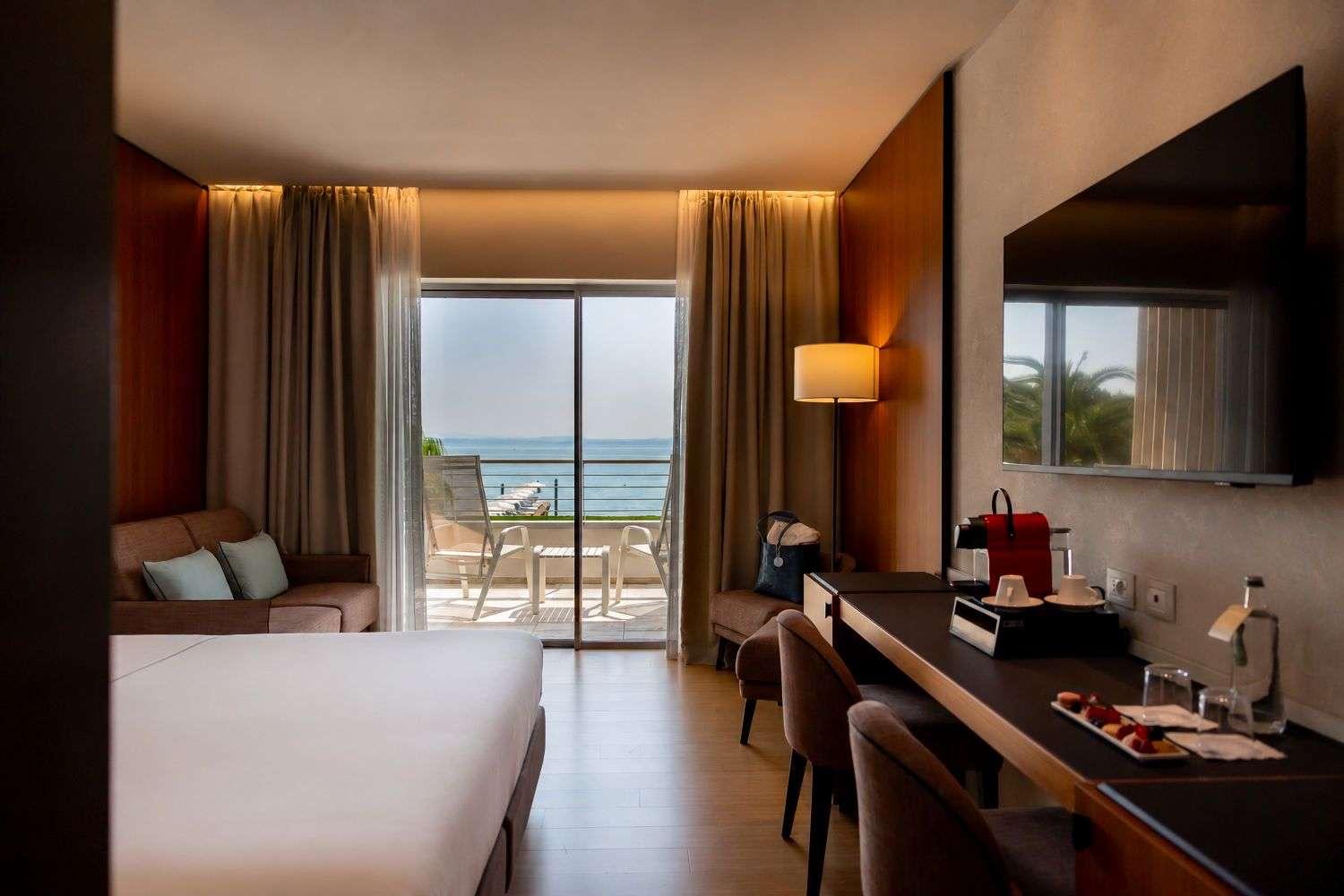 A hotel room with a bed, a sofa, and a breakfast tray on a counter, opening onto a balcony with lake views.