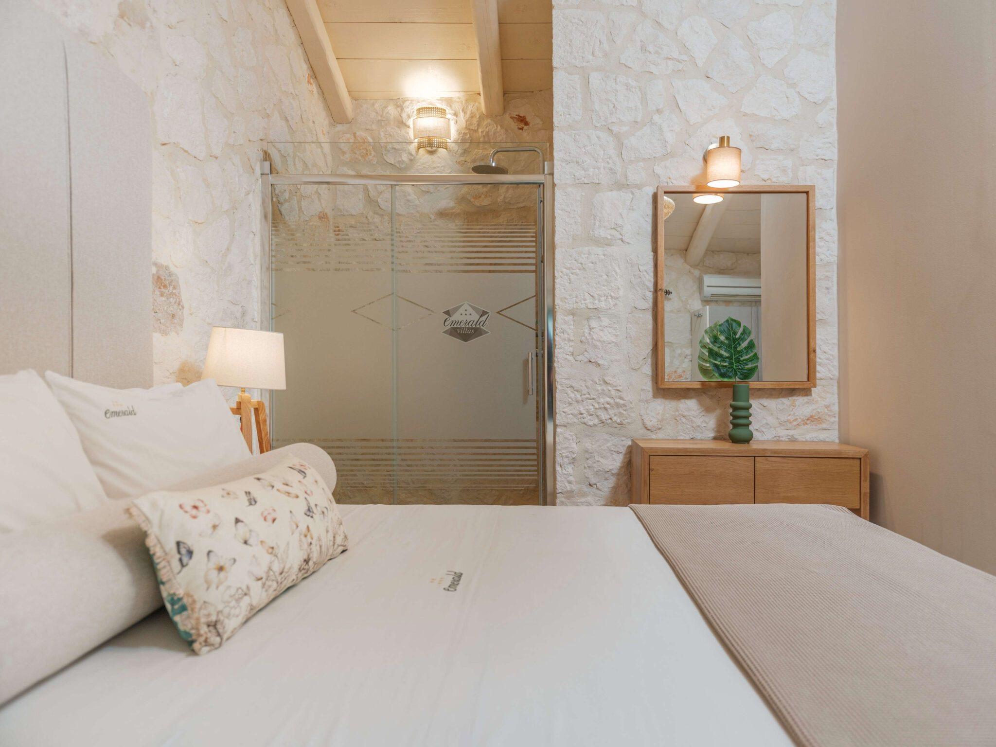 a bed with a monogrammed pillow against a stone wall, facing a glass shower door etched with the hotel's logo and a mirror reflecting a potted plant