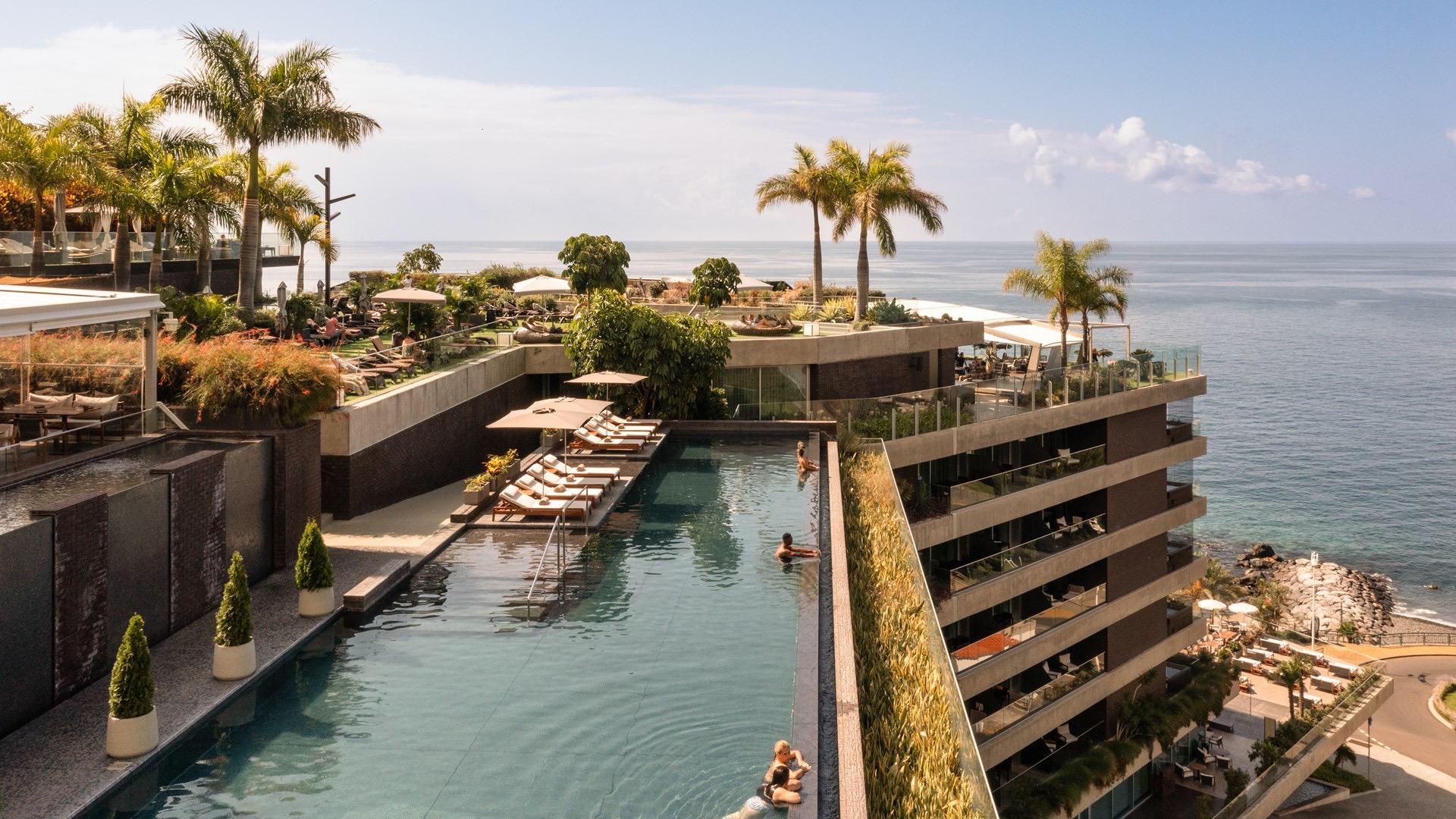 A rooftop infinity pool lined with palm trees and sun loungers, with two swimmers in the water and the ocean beyond.