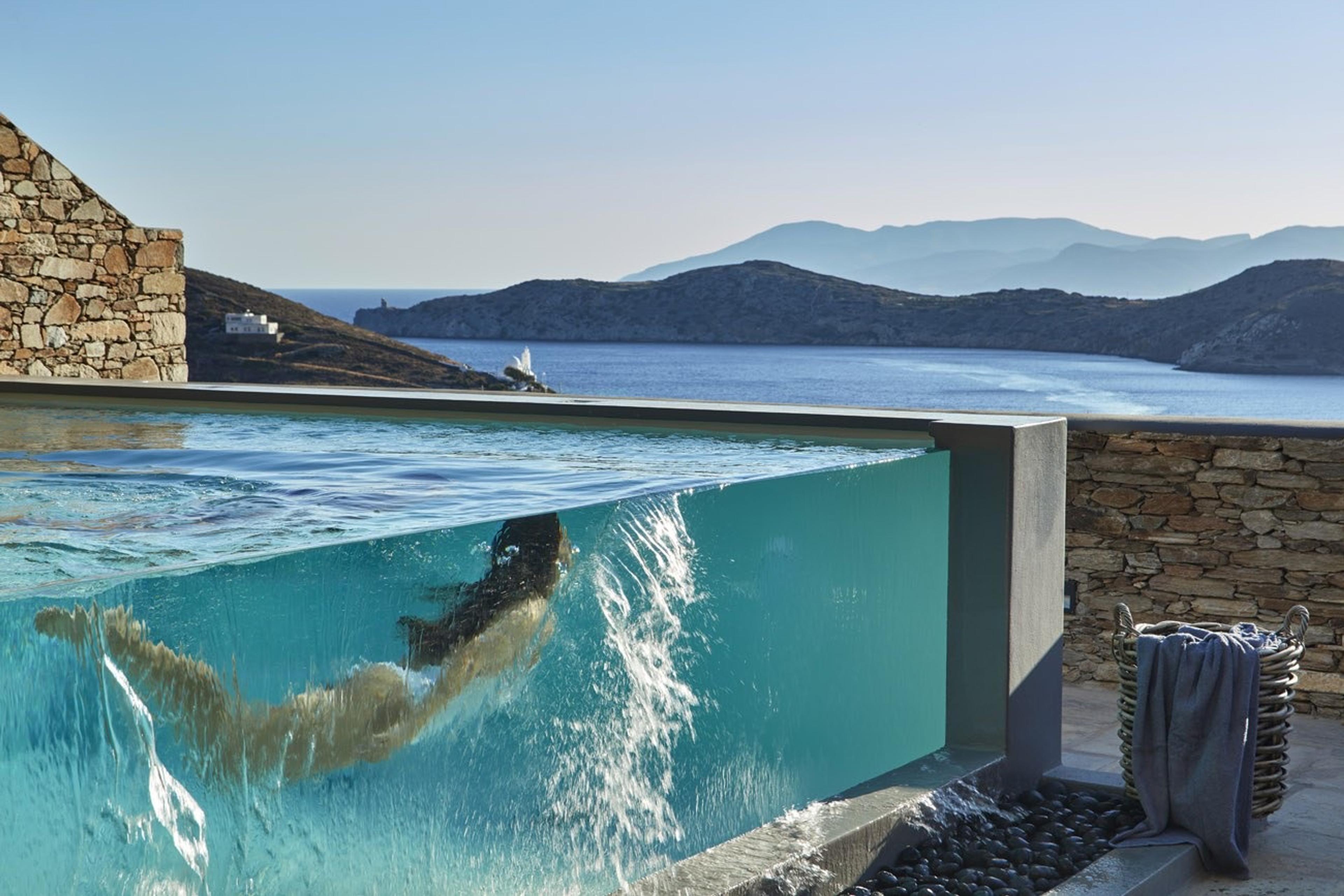 An infinity-edge pool sits above the sea, its glass side showing a swimmer's blurred shape underwater, with a stone wall and a folded towel in a basket beside it.