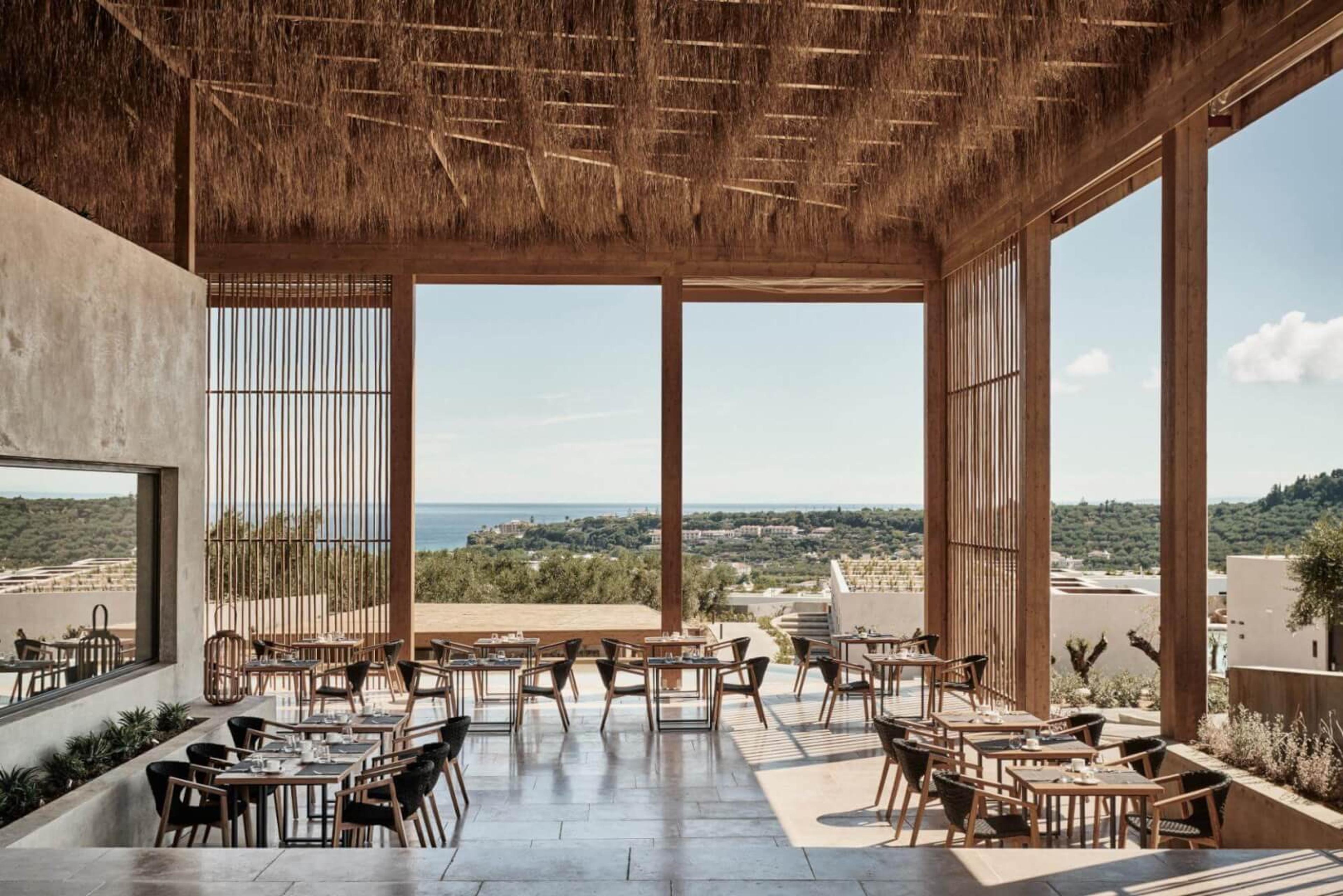 An open-sided restaurant terrace with a thatched roof, dark wood tables laid for a meal, and the sea visible between timber screens.