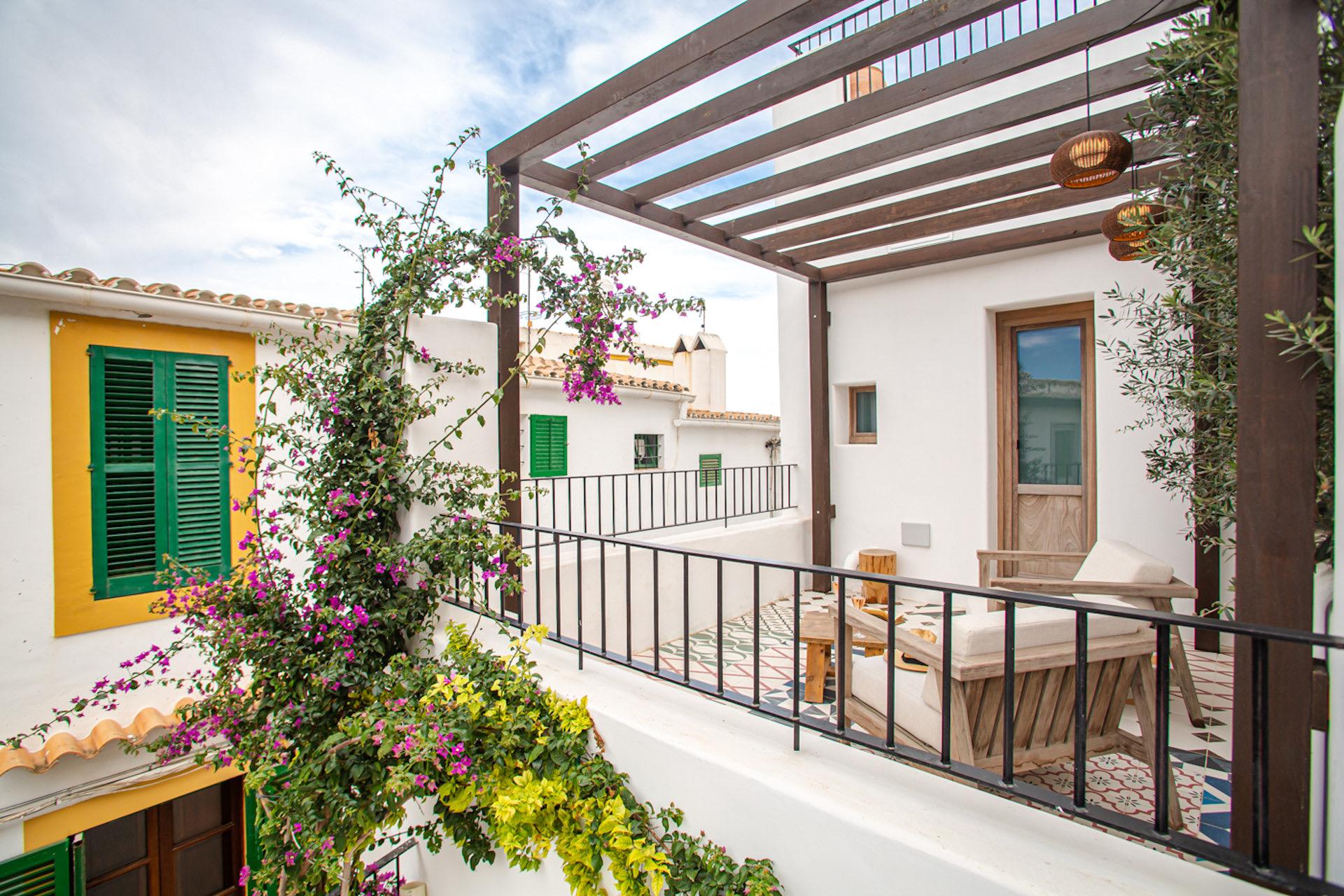 A first-floor terrace under a timber pergola with woven lamps, a bougainvillea climbing a white wall, and a wooden daybed with cushions beside a green-shuttered house.