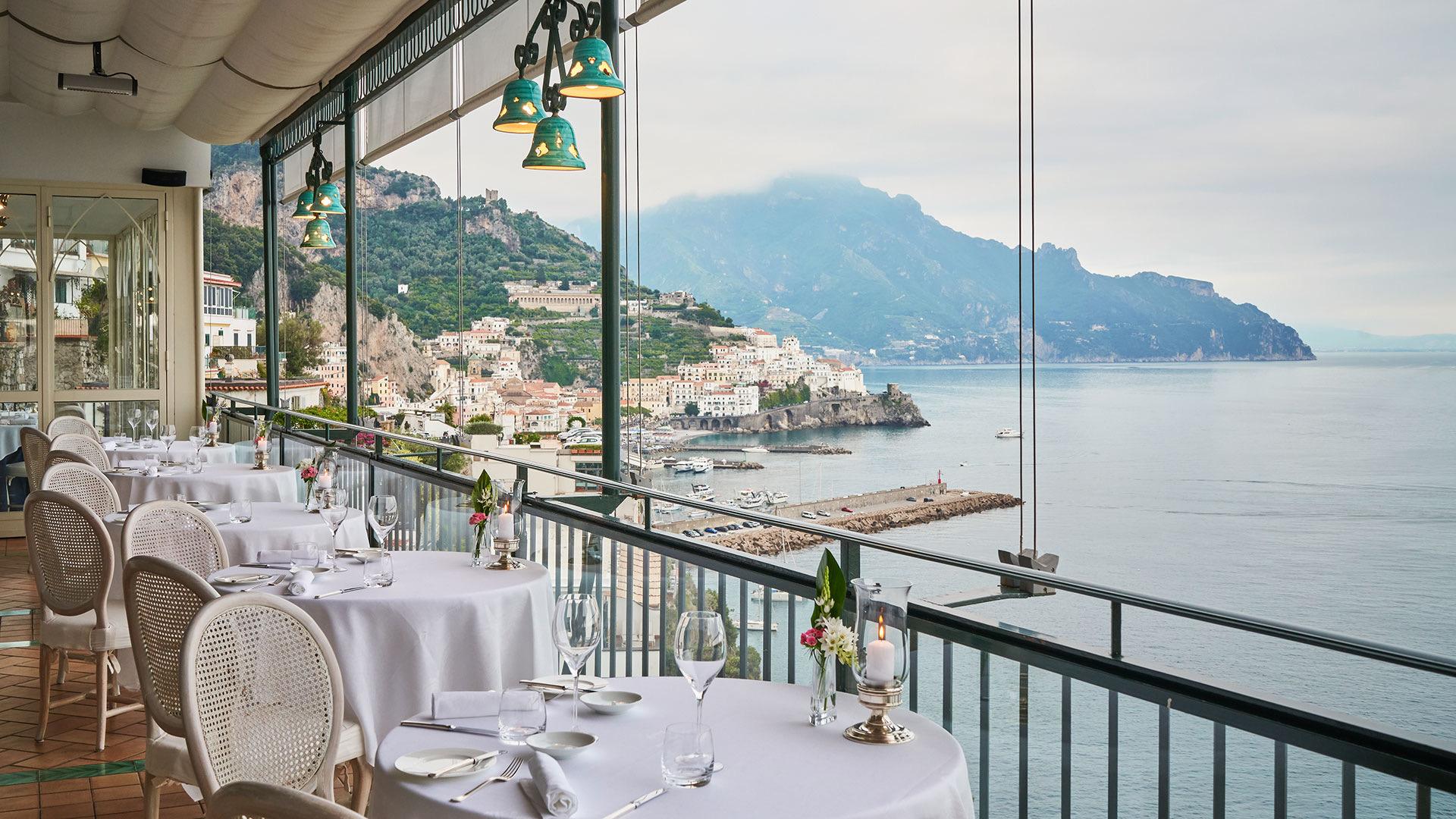 Tables laid in white along a glass-walled terrace, Amalfi's houses climbing the hill across the bay.