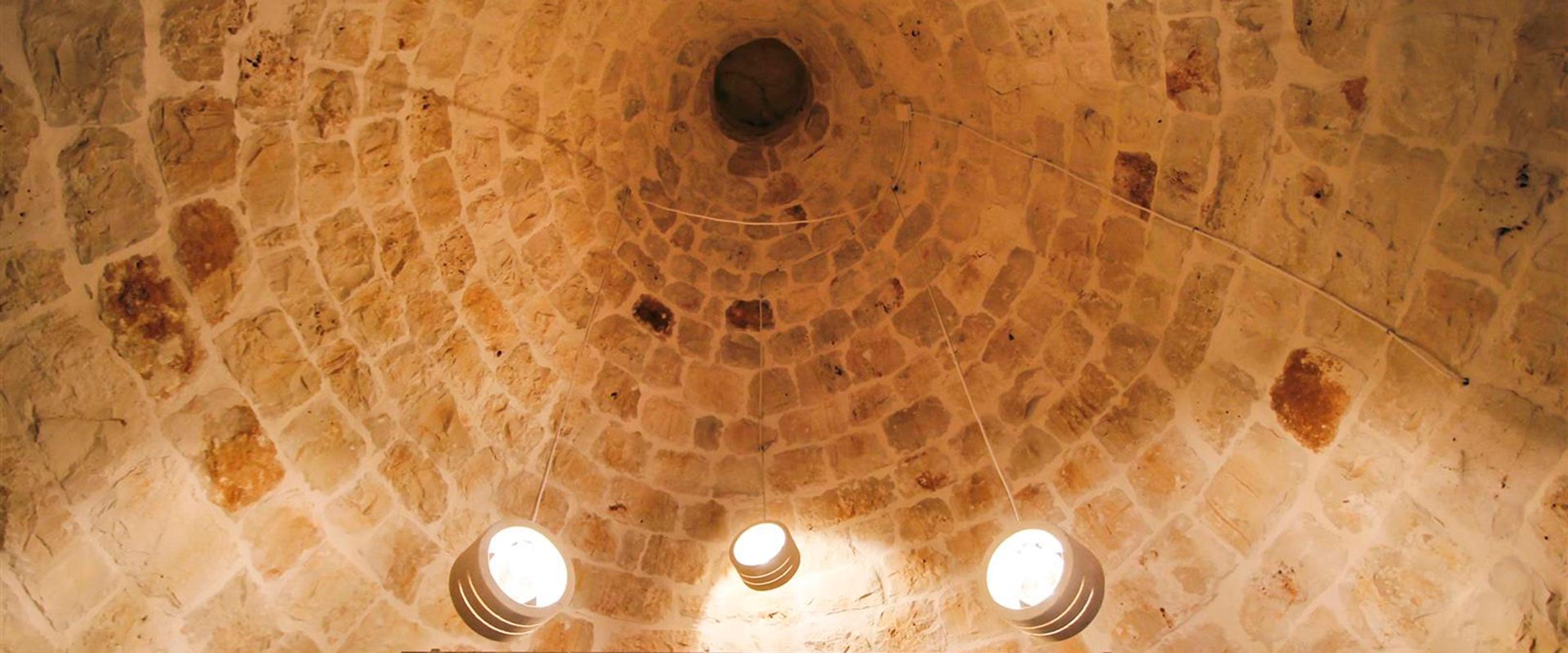 The inside of a conical stone trullo roof, its stones laid in rings up to a dark oculus, with pendant lights hanging below.