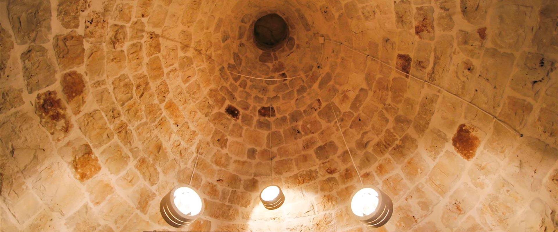 The inside of a conical stone trullo roof, its stones laid in rings up to a dark oculus, with pendant lights hanging below.