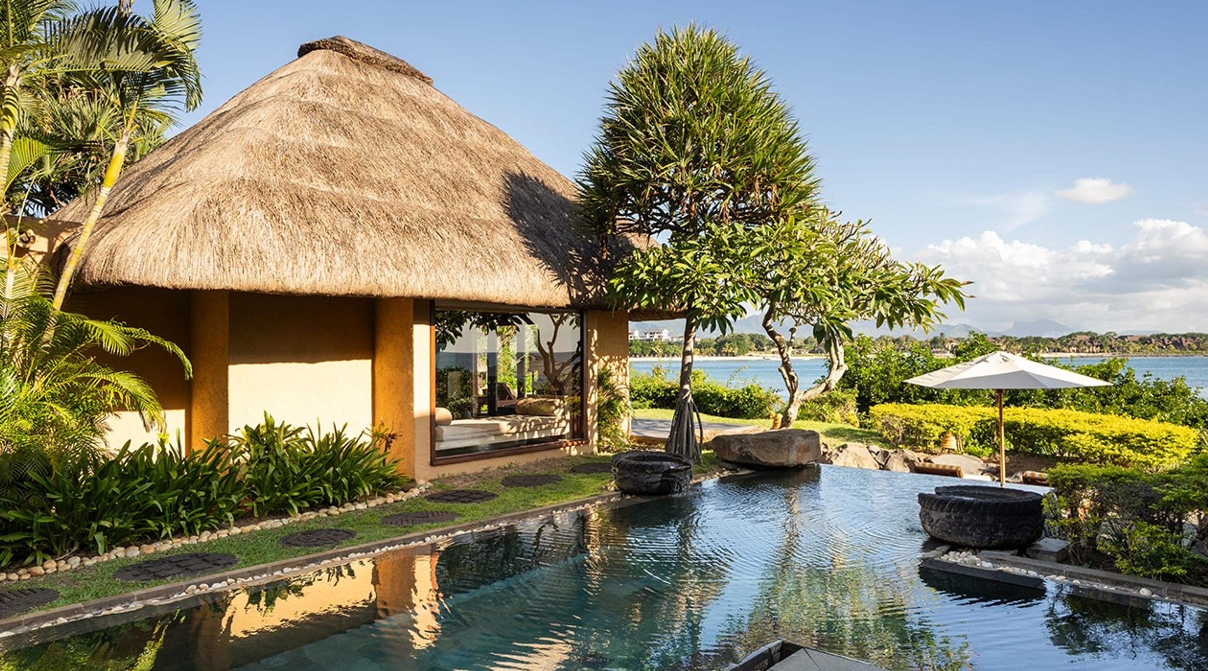 A thatched-roof villa beside an infinity pool that appears to merge with the sea, with an umbrella and hills visible across the water.