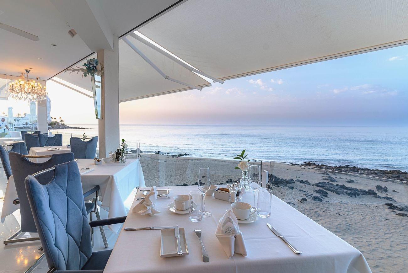 An empty restaurant terrace with blue upholstered chairs and tables set for dinner, under a canopy with the sea and a sandy shore beyond.