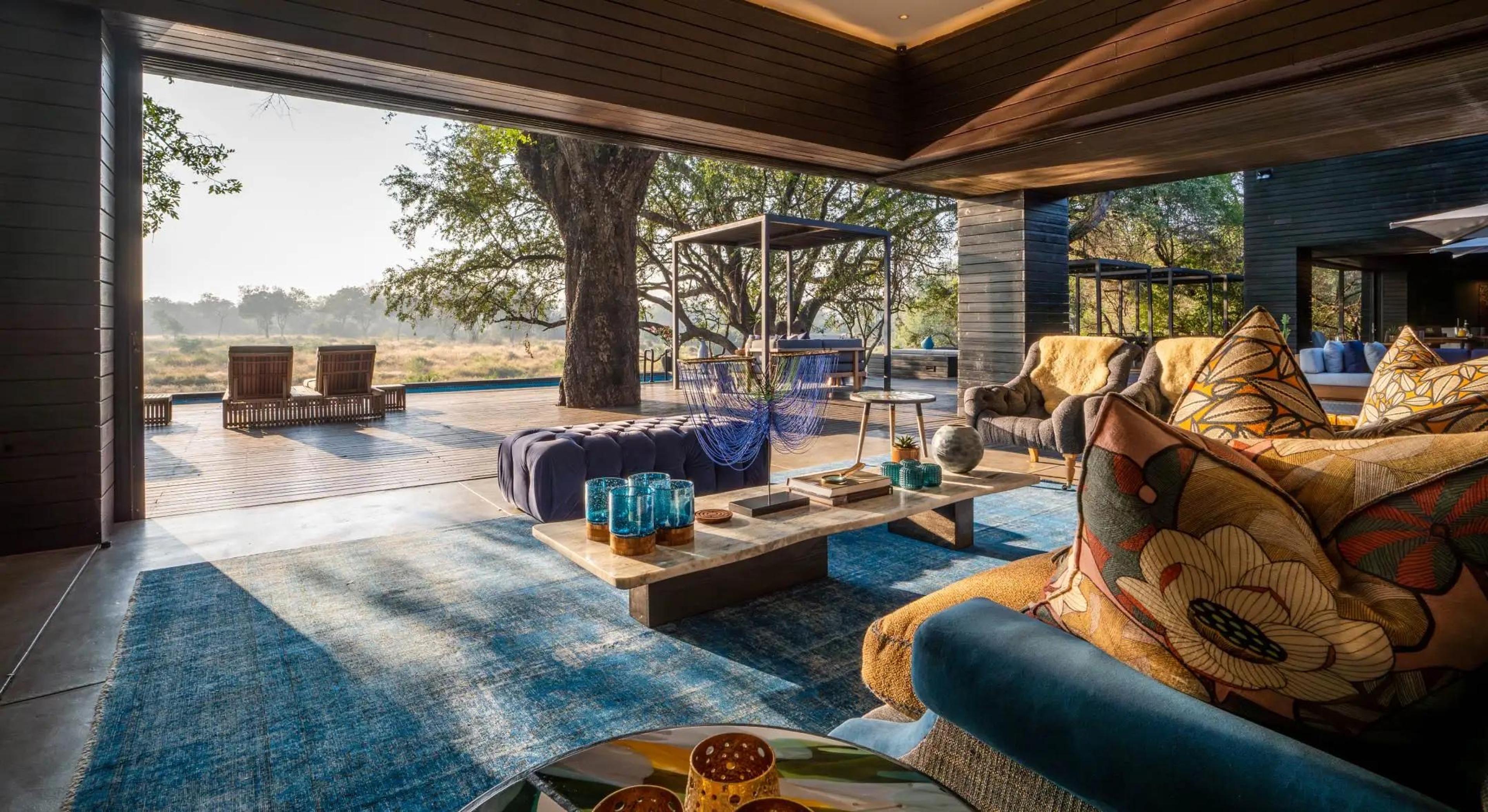 An open-sided lounge with a blue rug and dark blue sofas, looking through a wide opening onto a wooden deck with sun loungers, a large tree, and grassland beyond.