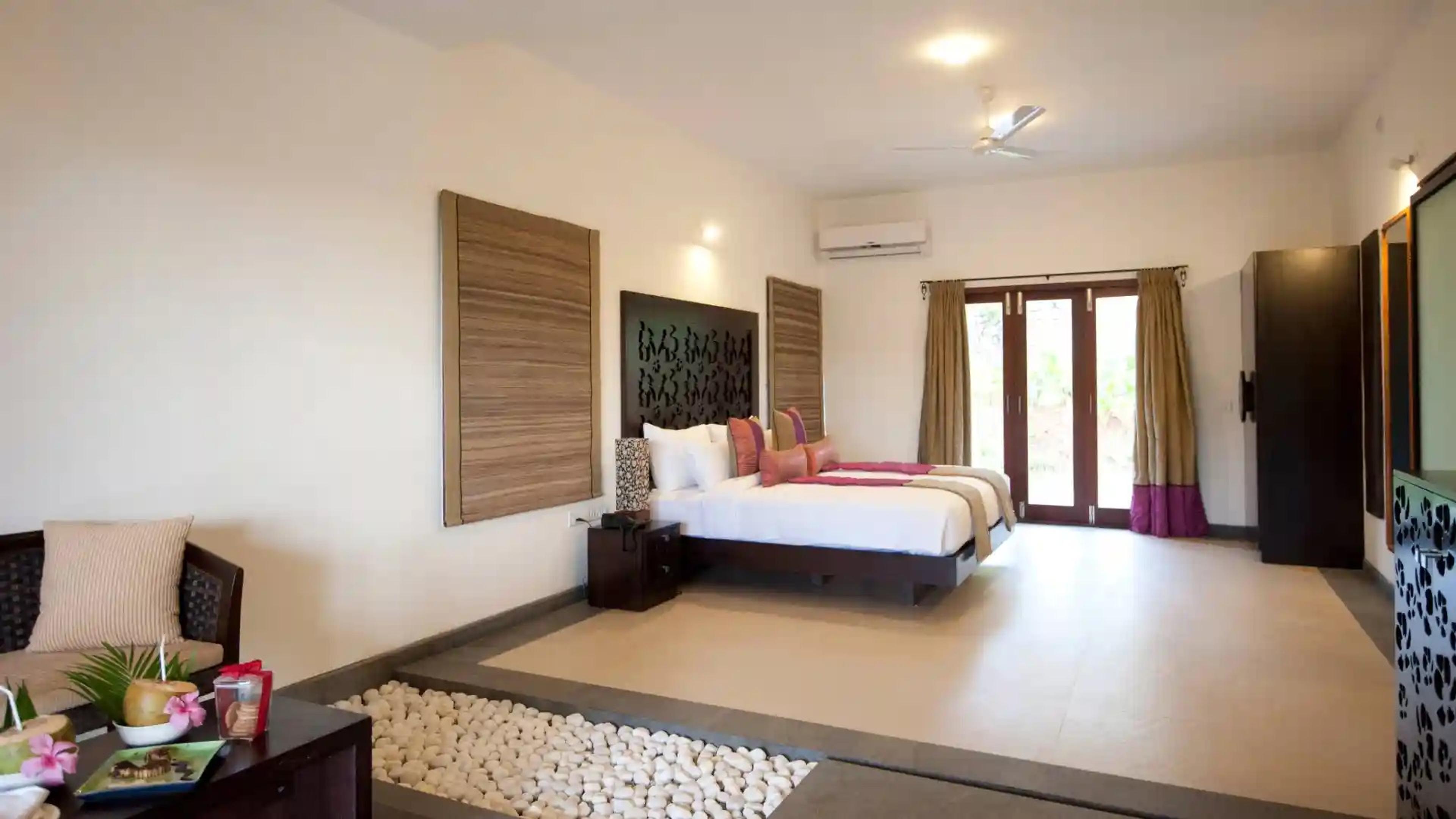 a bedroom with a dark wood bed, woven wall panels and glass doors open to a garden, with a seating area and pebble path