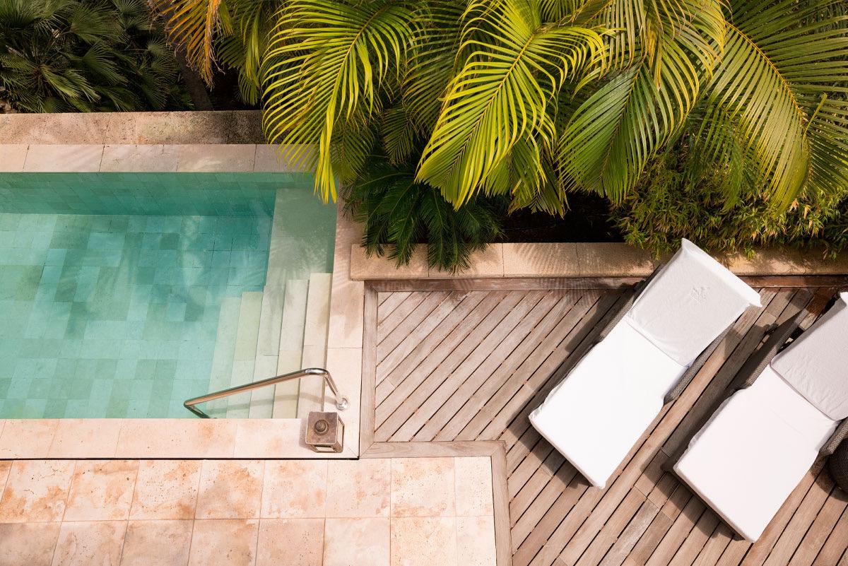An overhead view of a small plunge pool and a wooden sun deck with two loungers, bordered by palm fronds.