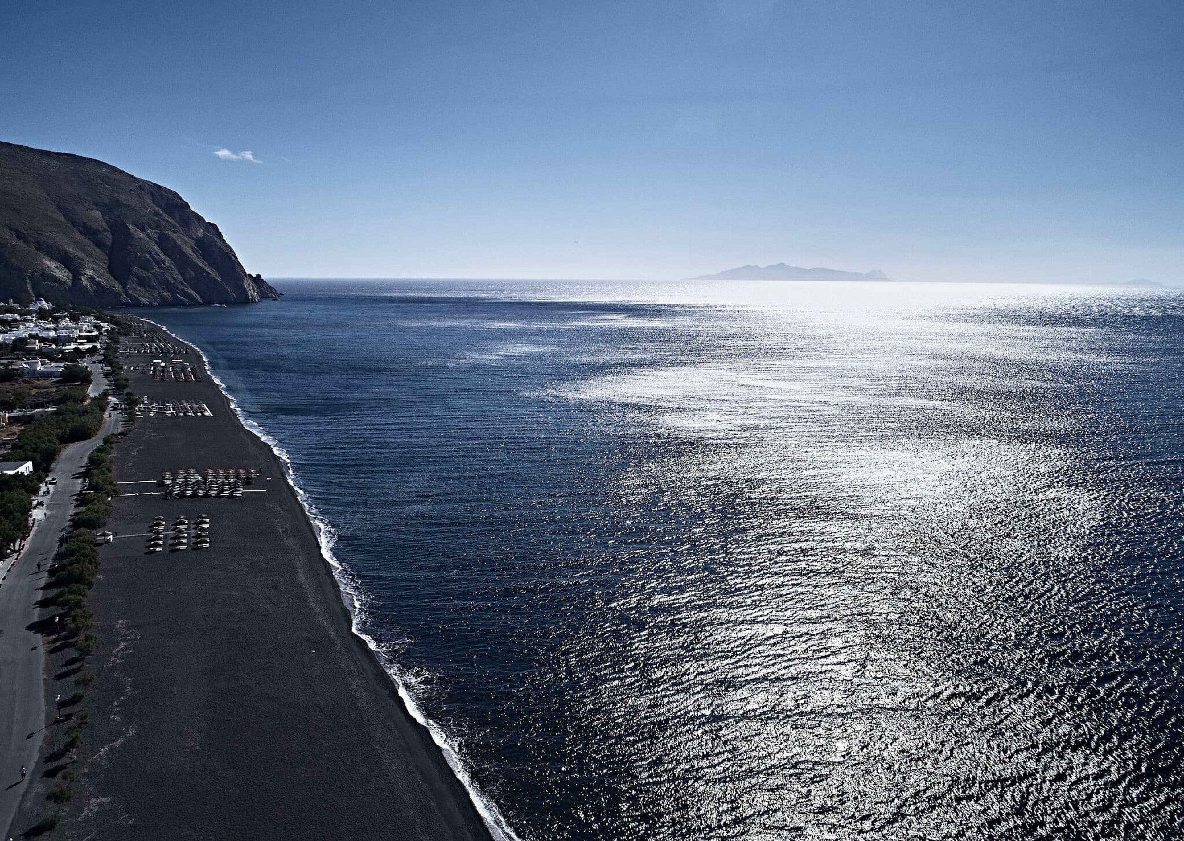 An aerial view of a black sand beach along a headland, with sunbeds under the cliff and the sea stretching to the horizon.