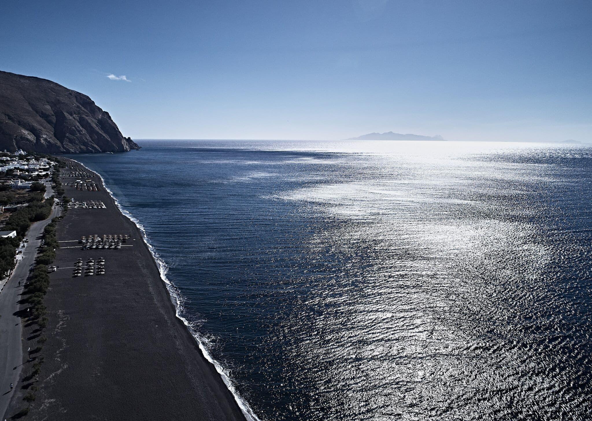 An aerial view of a black sand beach along a headland, with sunbeds under the cliff and the sea stretching to the horizon.