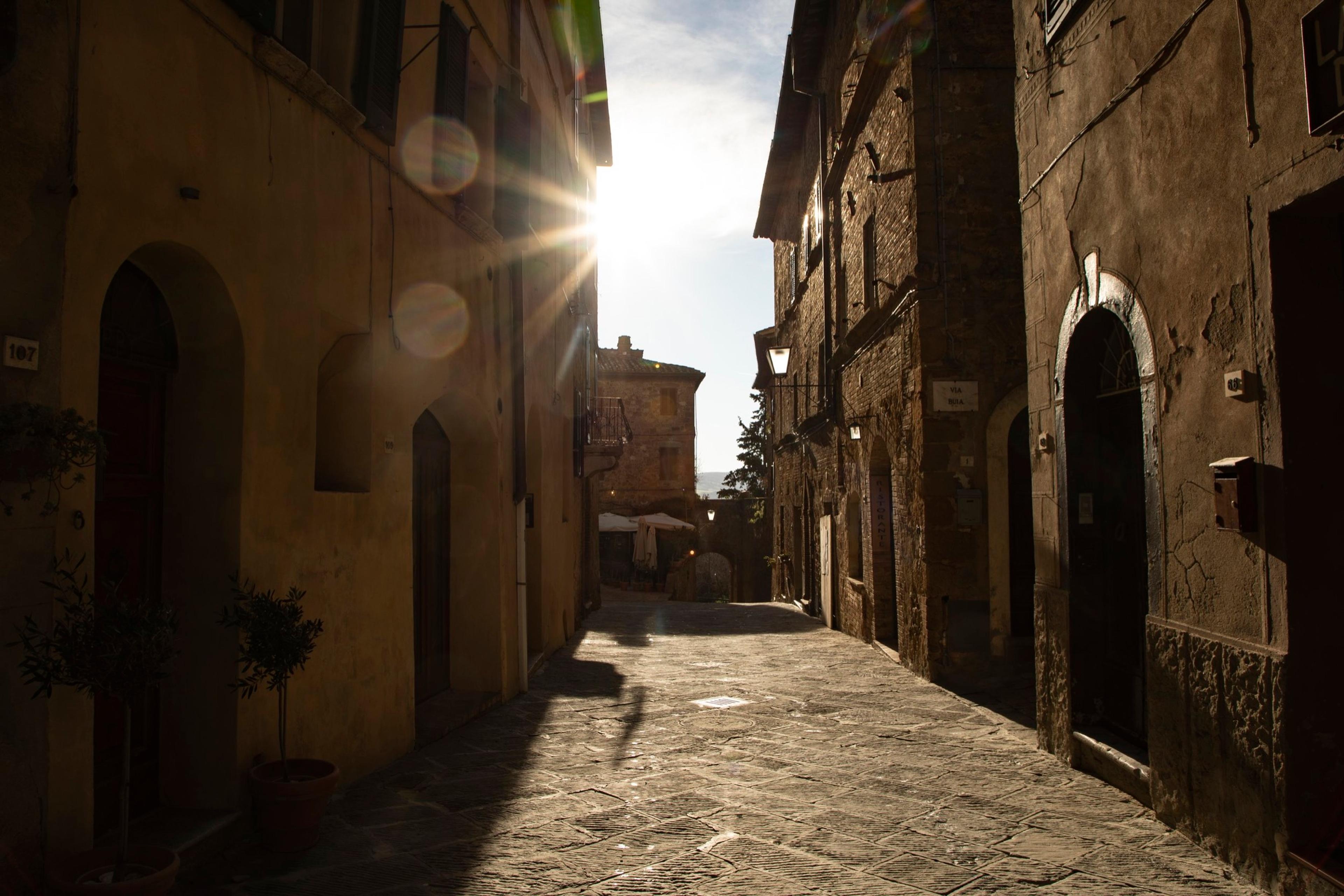 A narrow stone alley between tall buildings, lit by low sun flaring over the rooftops.