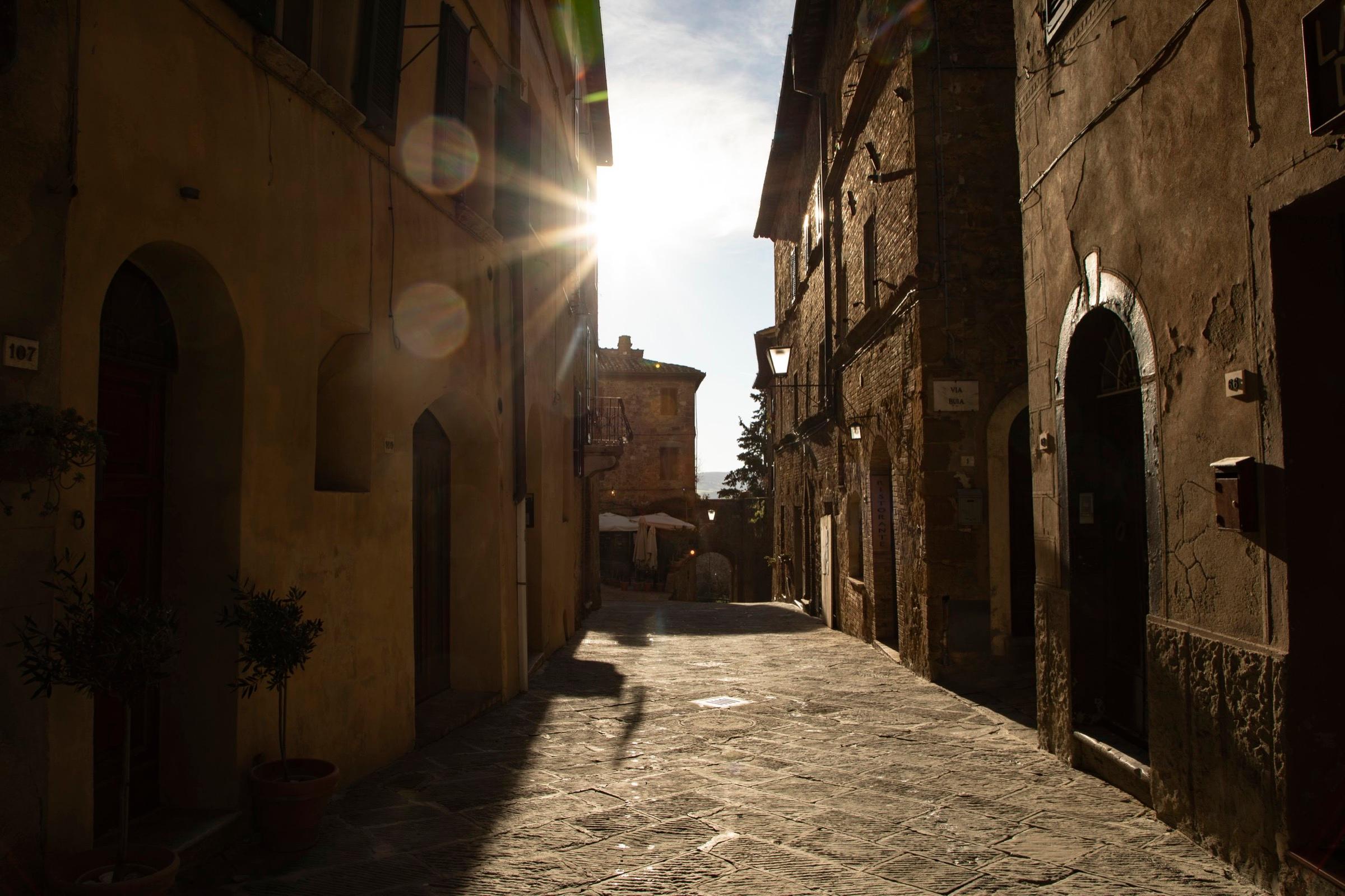 A narrow stone alley between tall buildings, lit by low sun flaring over the rooftops.