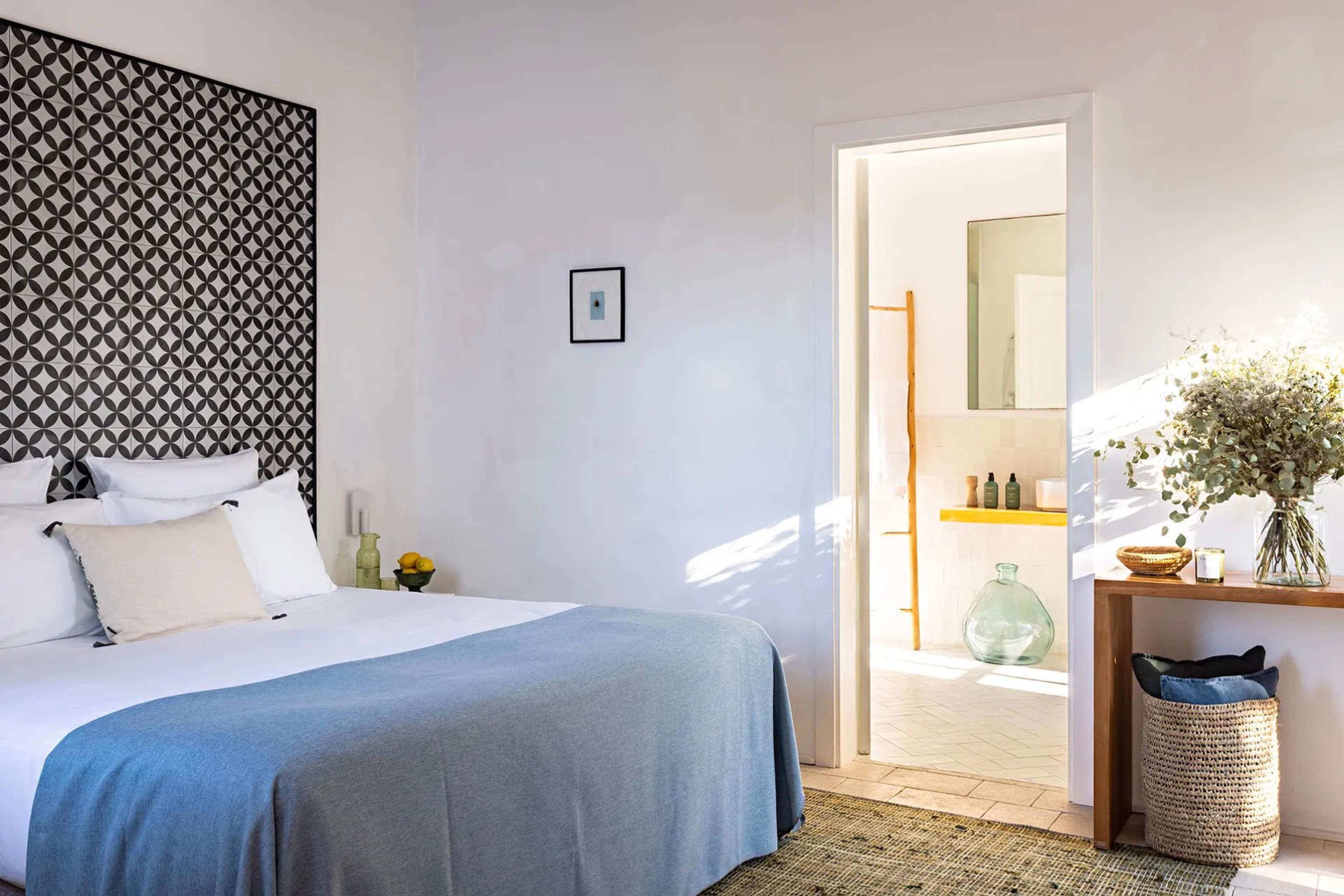 a bedroom with a black and white mosaic tiled headboard and a blue throw, its door open onto an ensuite bathroom with a roll of towels and a glass jar