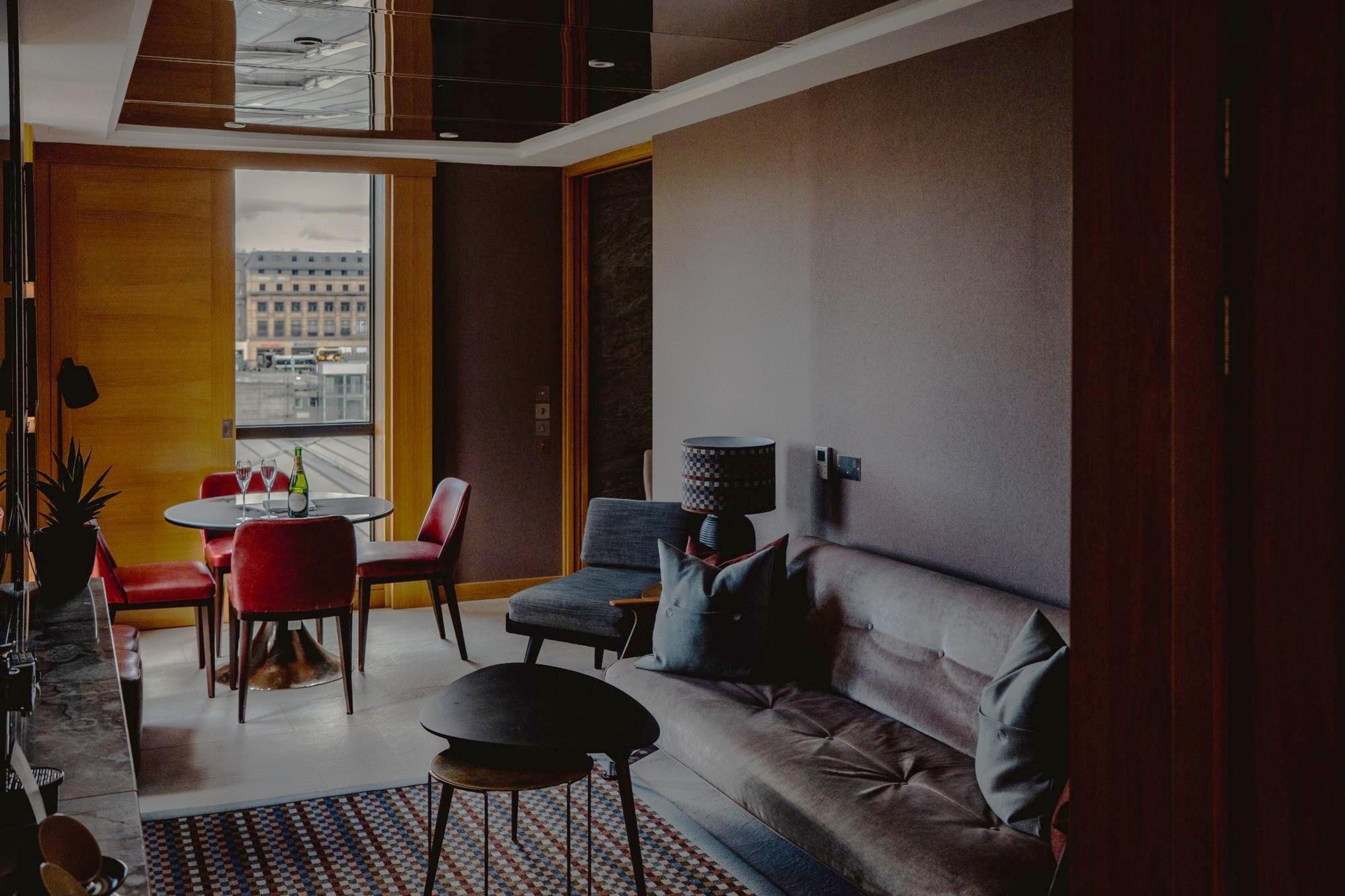 A suite sitting area with a grey sofa, a patterned rug, nested black coffee tables and a dining table with four red chairs.