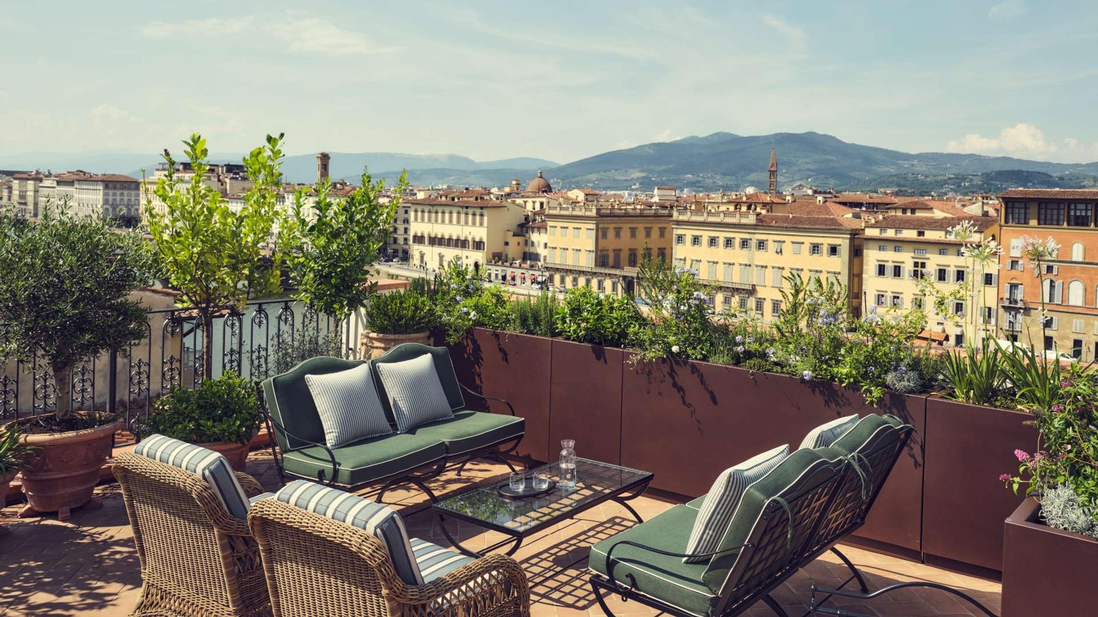 A rooftop terrace with wicker chairs and green cushions around a low table, potted olive trees at the railing, looking out over Florence's rooftops to the hills beyond.