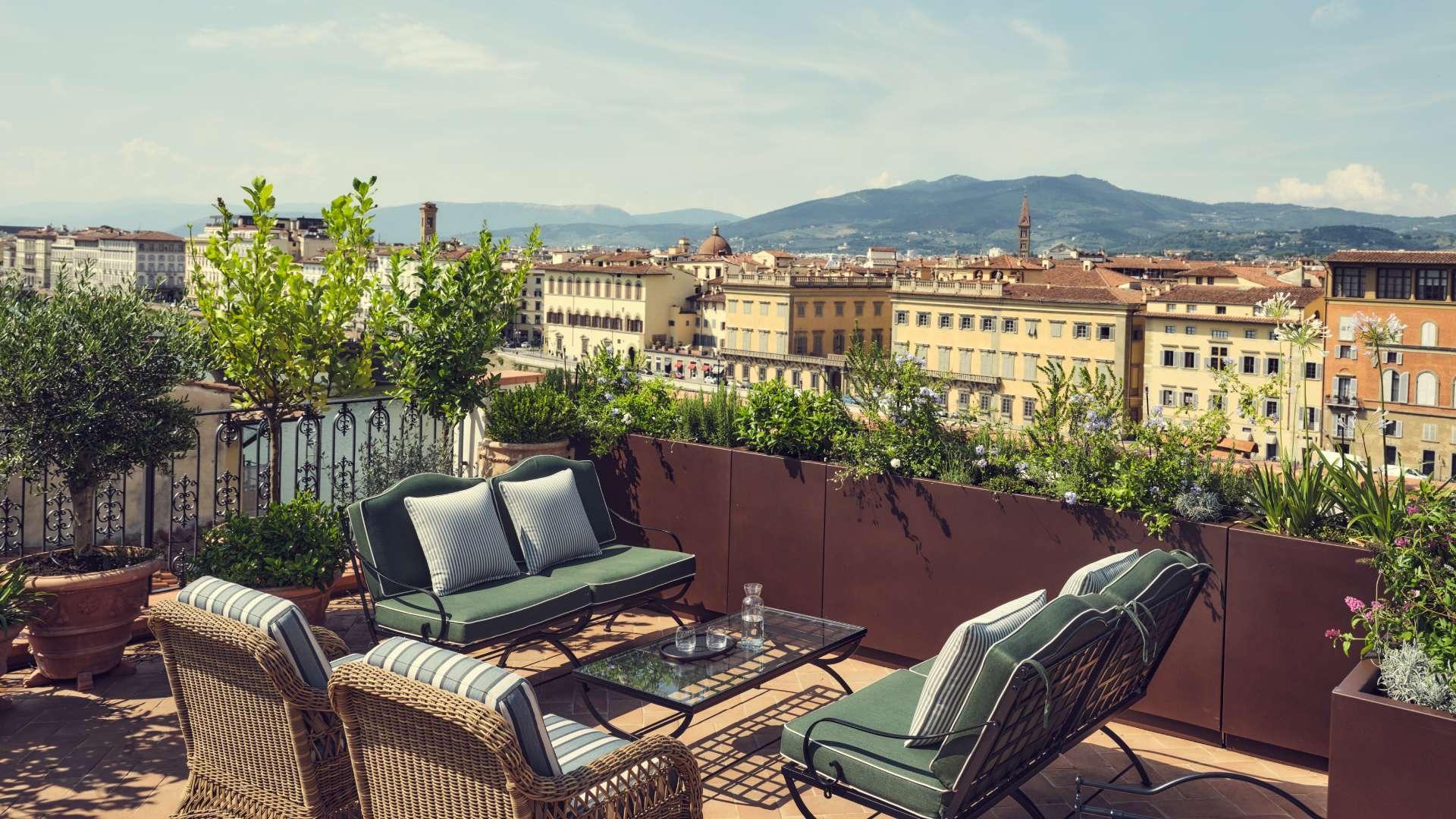 A rooftop terrace with wicker chairs and green cushions around a low table, potted olive trees at the railing, looking out over Florence's rooftops to the hills beyond.