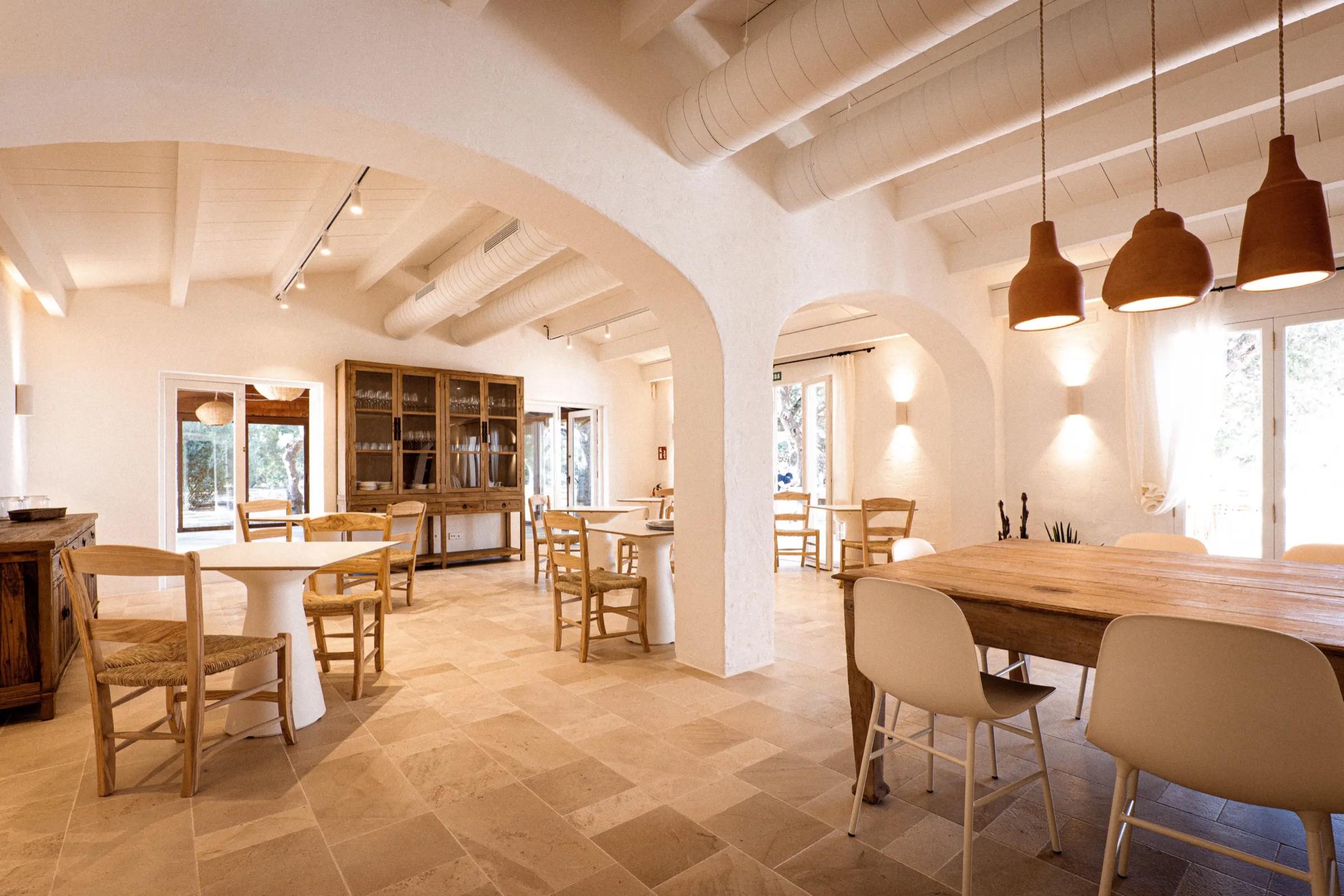 A whitewashed dining room under arches, wooden tables and chairs and woven pendant lamps.