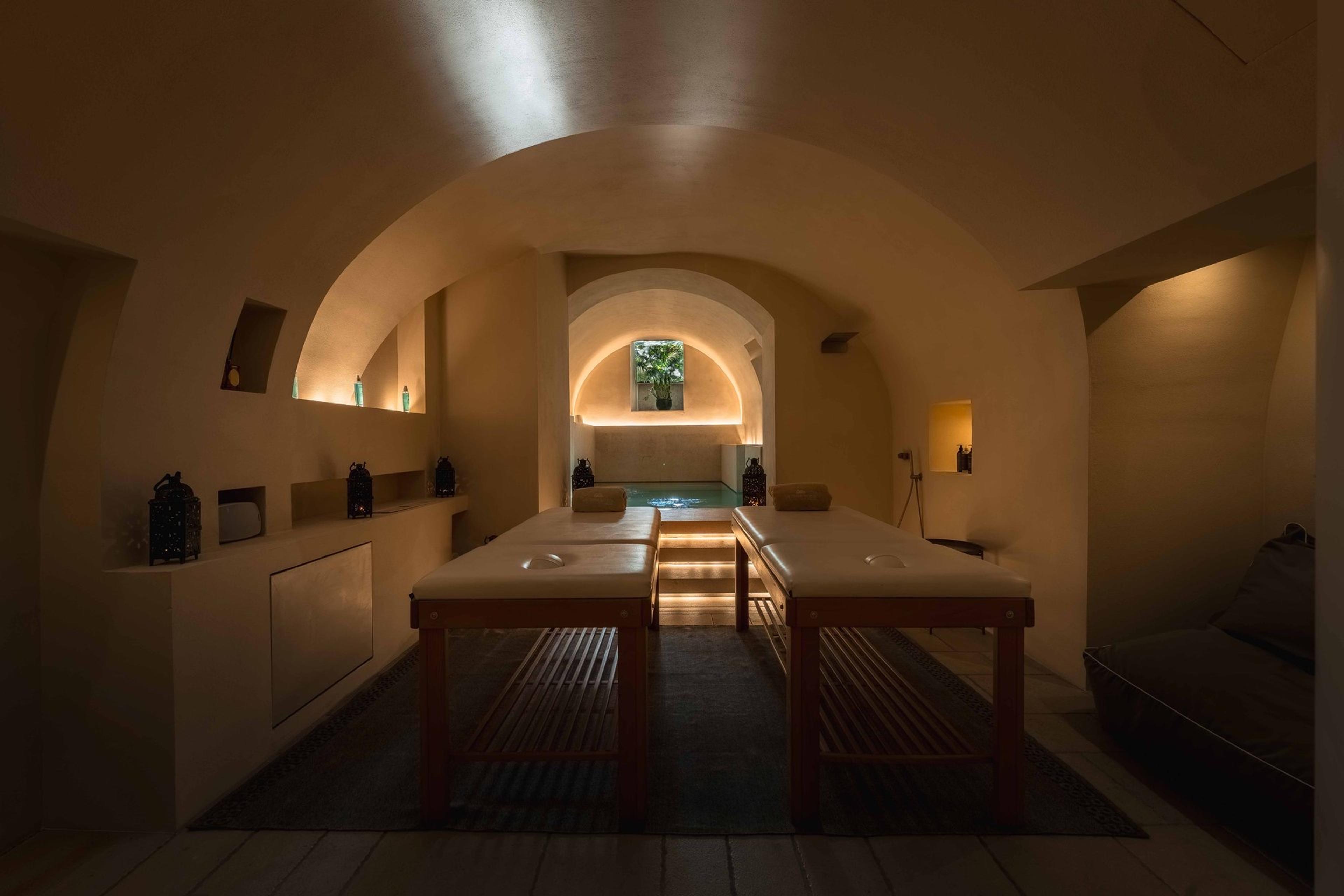 A dim room with two massage tables facing a small lit plunge pool under a vaulted stone ceiling.