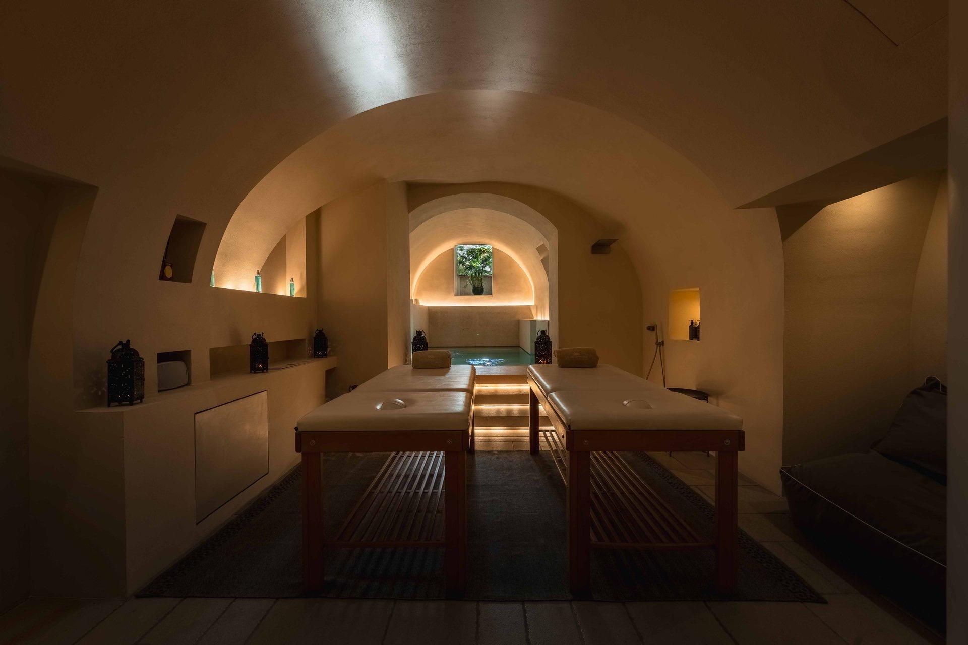A dim room with two massage tables facing a small lit plunge pool under a vaulted stone ceiling.