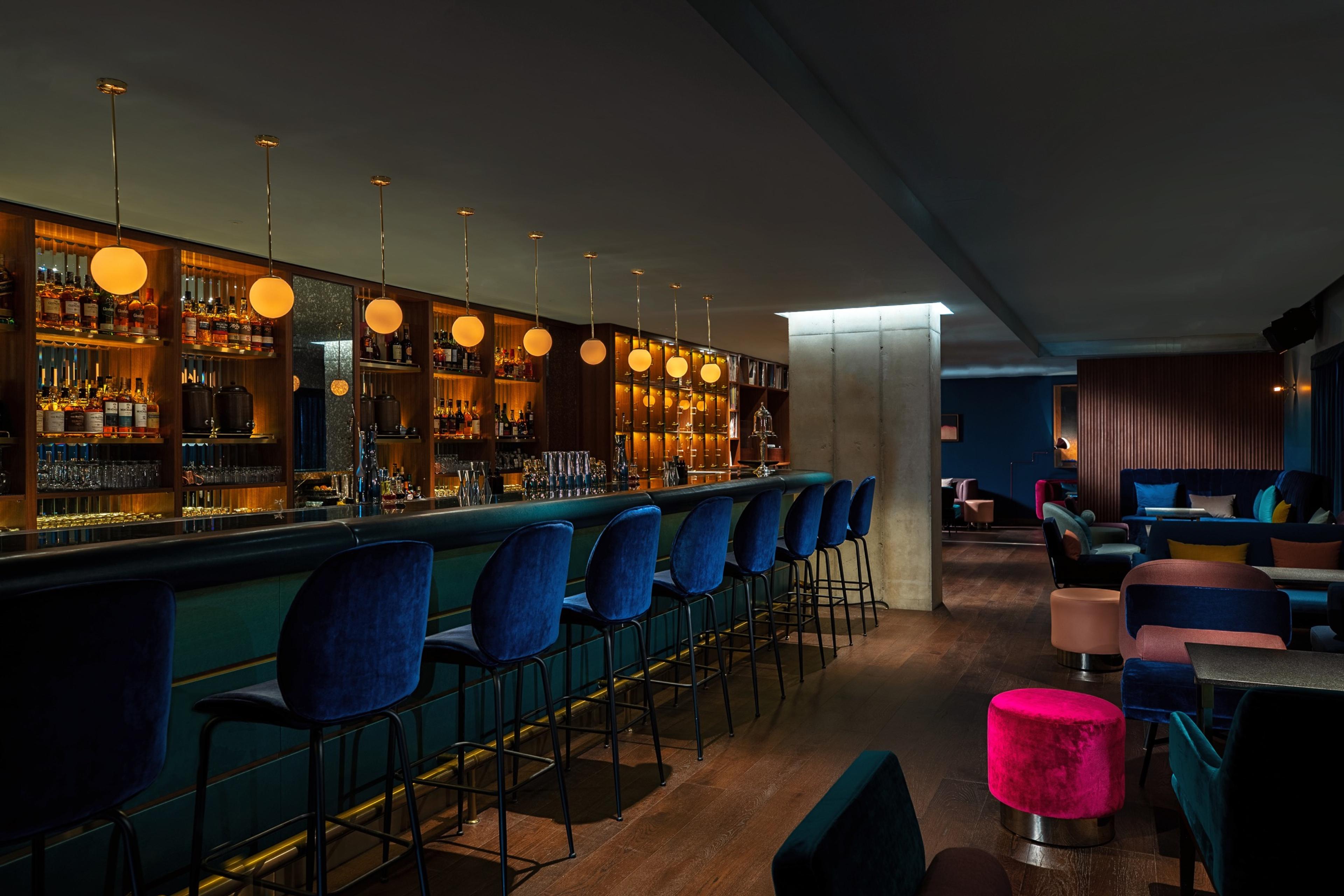 A long hotel bar with blue velvet stools and back-lit bottles on wood shelving, with pink and blue stools in the lounge beyond.