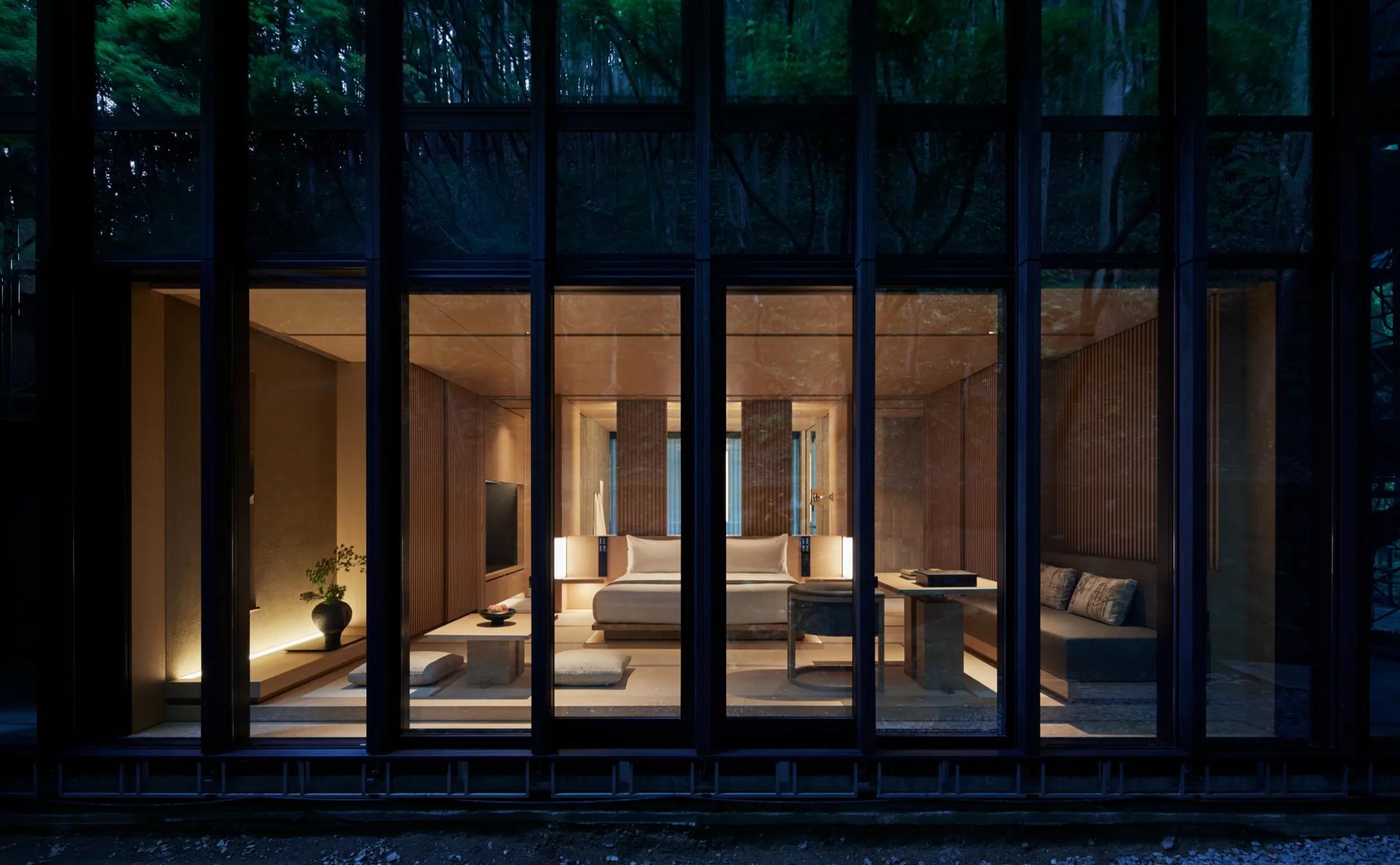 A guest room seen from outside at dusk through a glass facade, its bed and seating area lit warmly against dark surrounding forest.