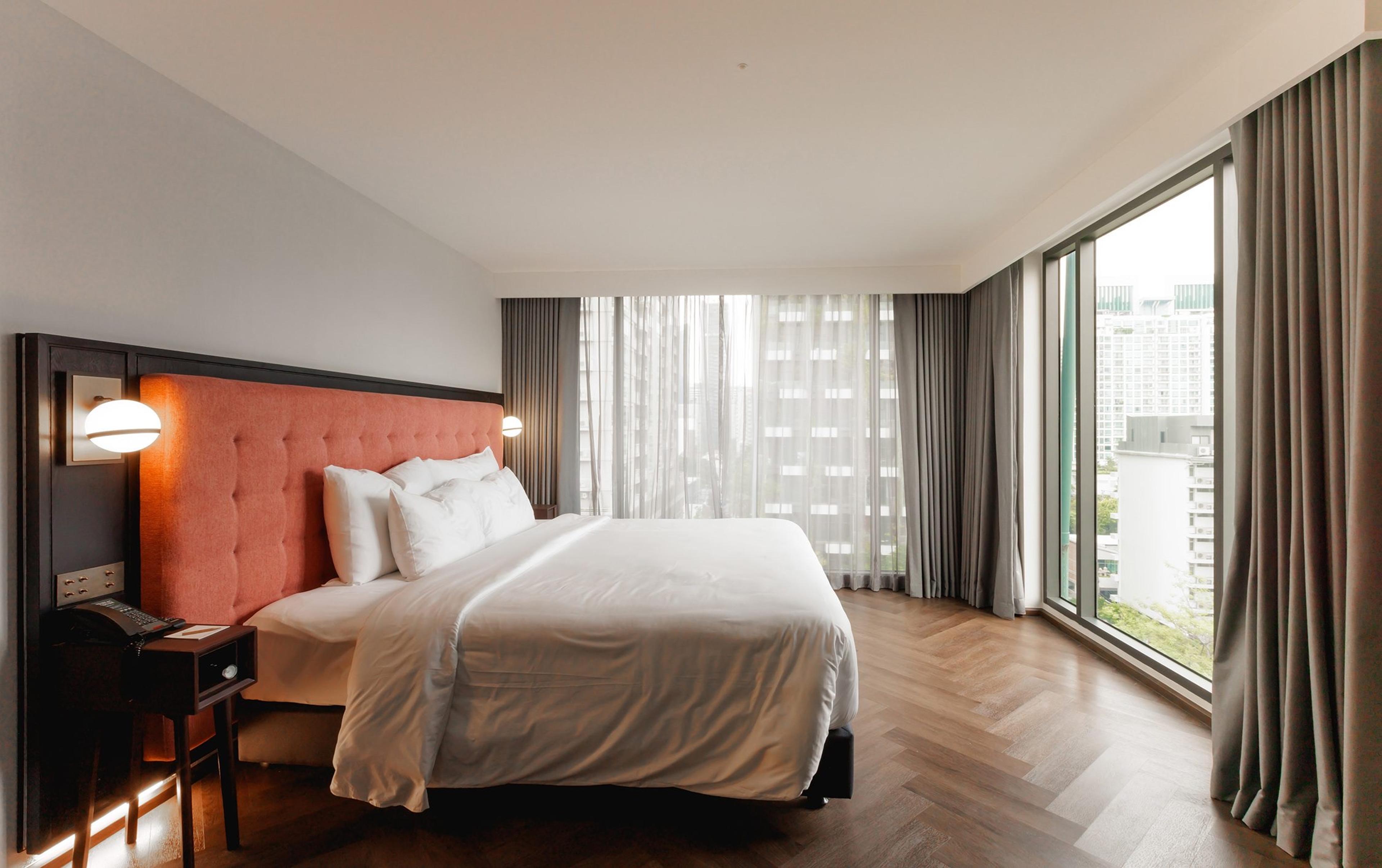 A bedroom with a coral upholstered headboard, a white bed, herringbone parquet floor and floor to ceiling windows on two walls showing the city outside.