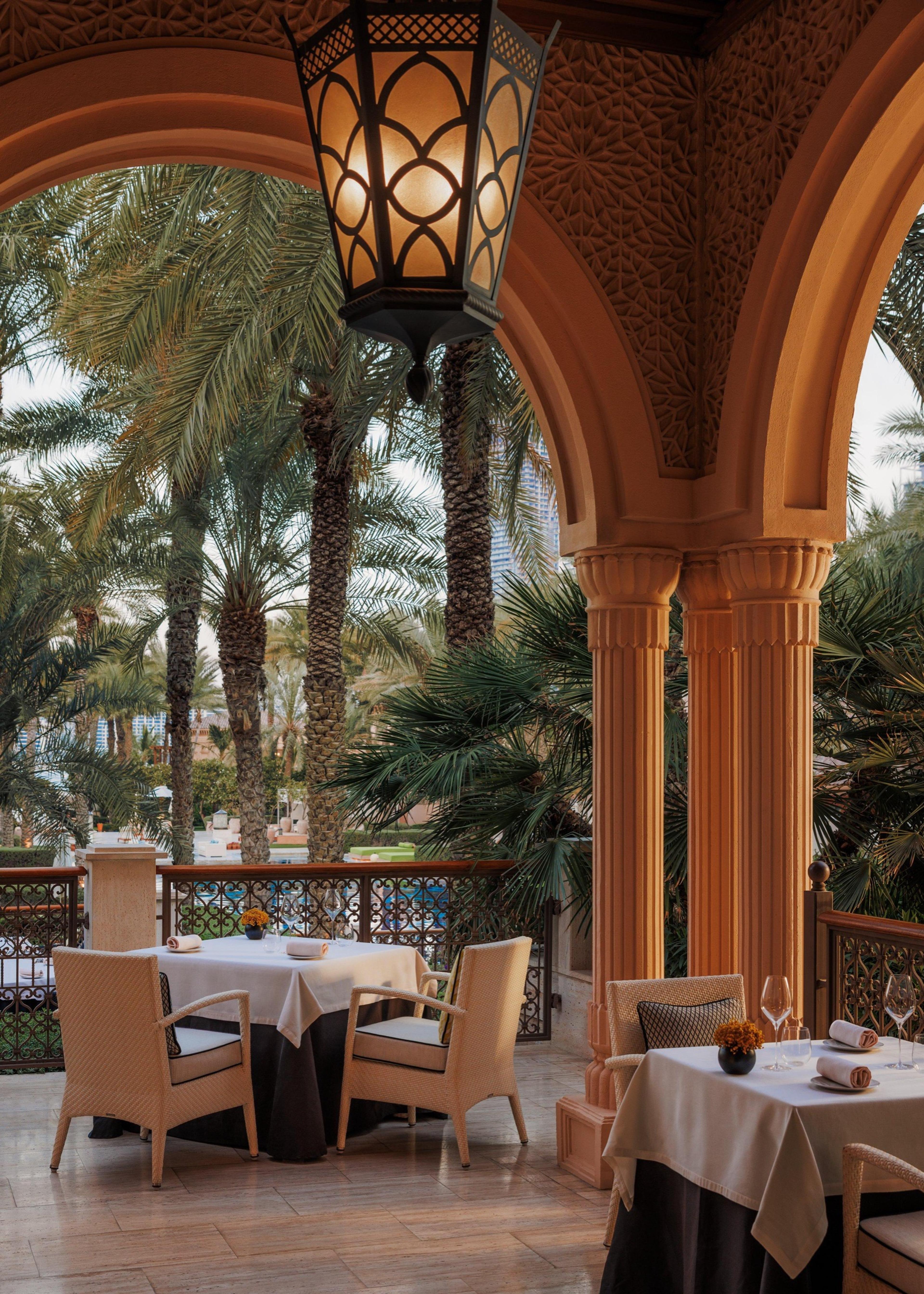 An empty restaurant terrace with arched columns and a lit lantern overlooks palm trees and a city skyline at dusk.