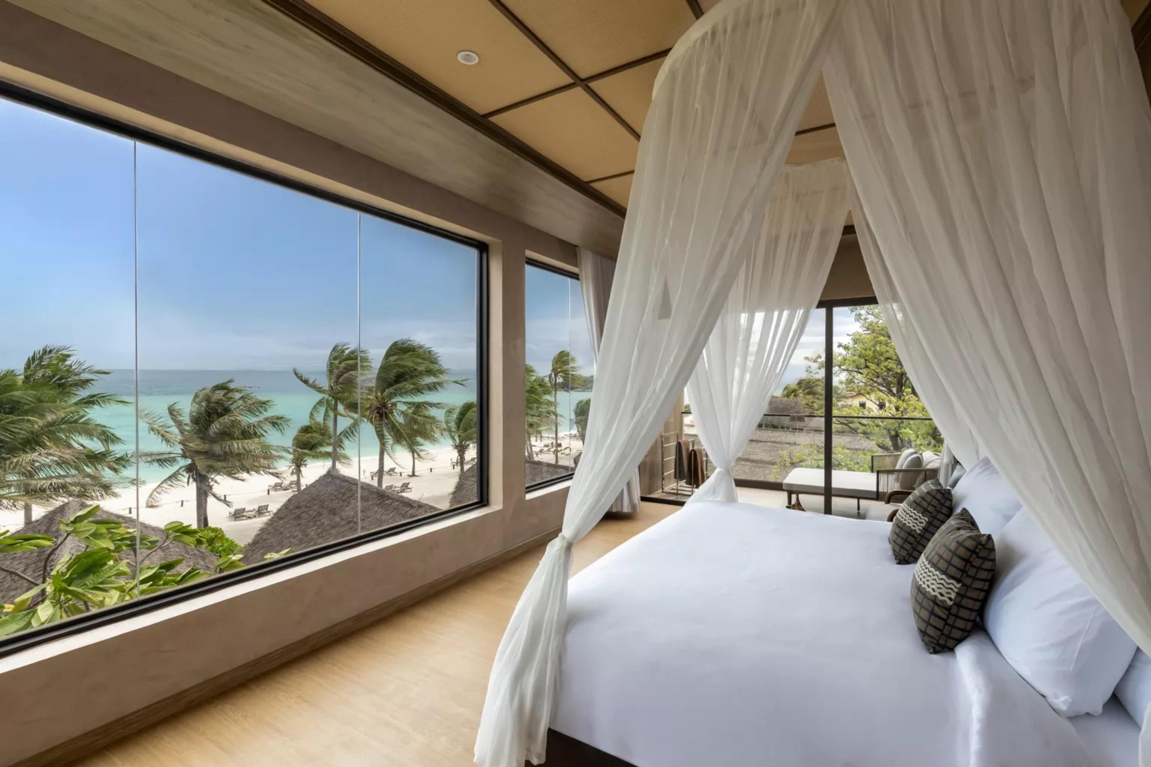 a canopy bed facing a wall of windows that frame a white sand beach, turquoise sea and palm trees bending in the wind
