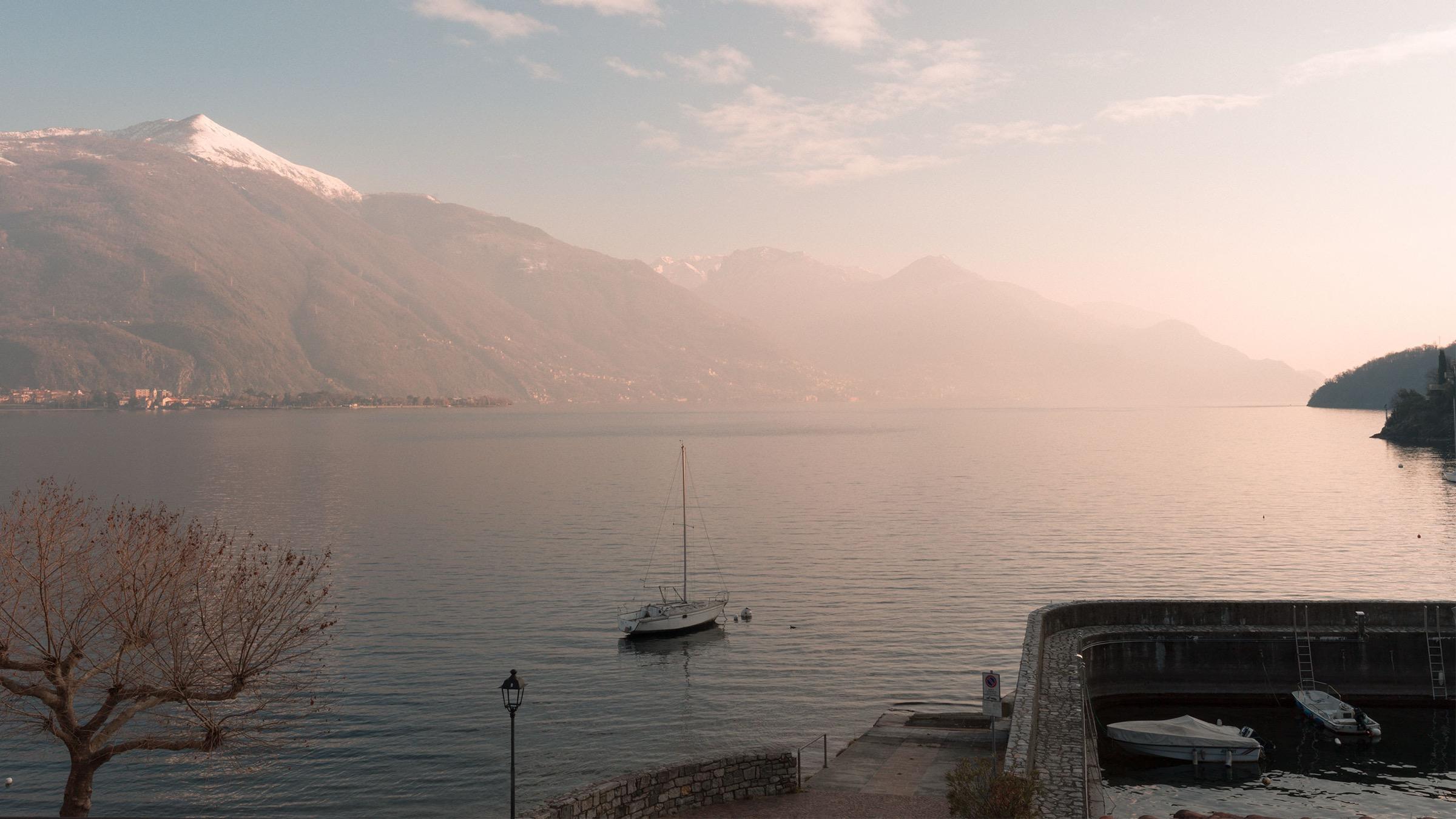 a wide lake view at dawn with snow capped mountains, a small boat, and a stone jetty with moored boats