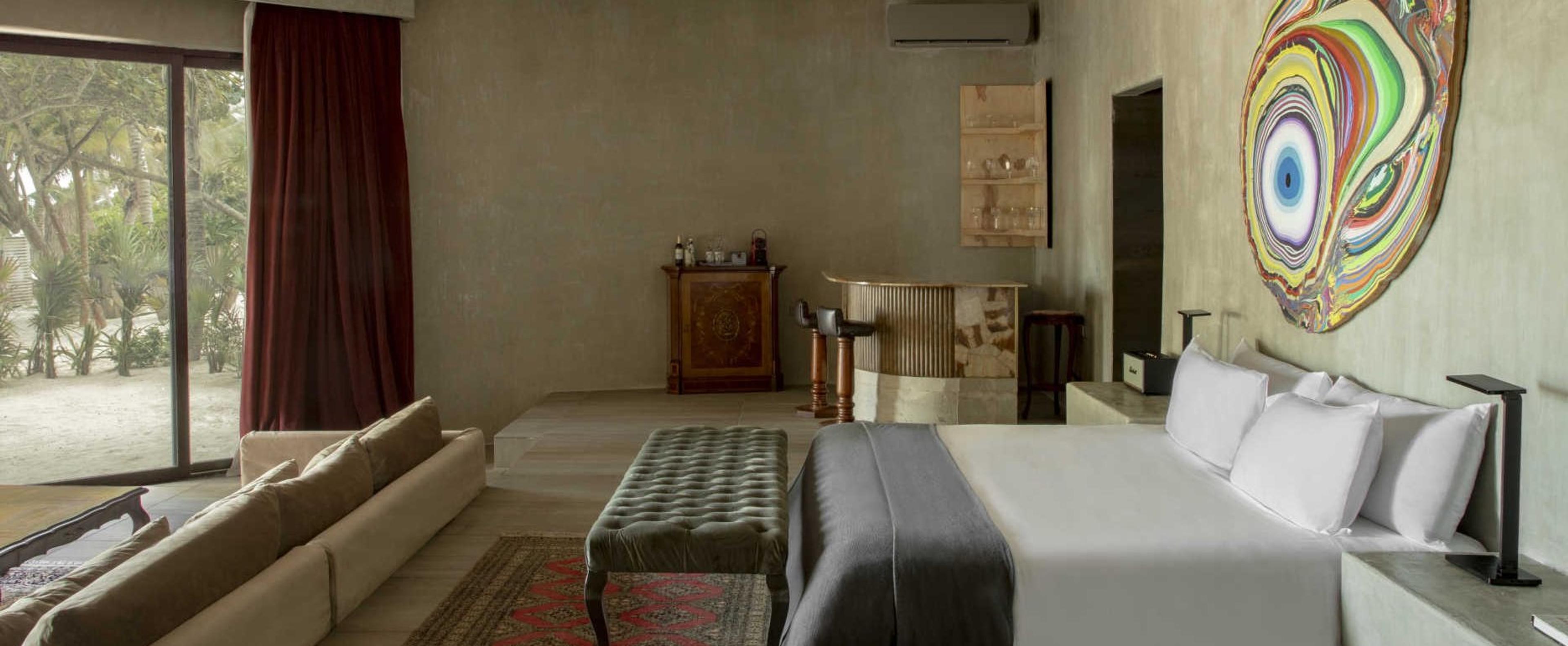 A hotel suite with a sofa, a tufted bench, a bed and a small bar, lit by a glass wall onto a garden.
