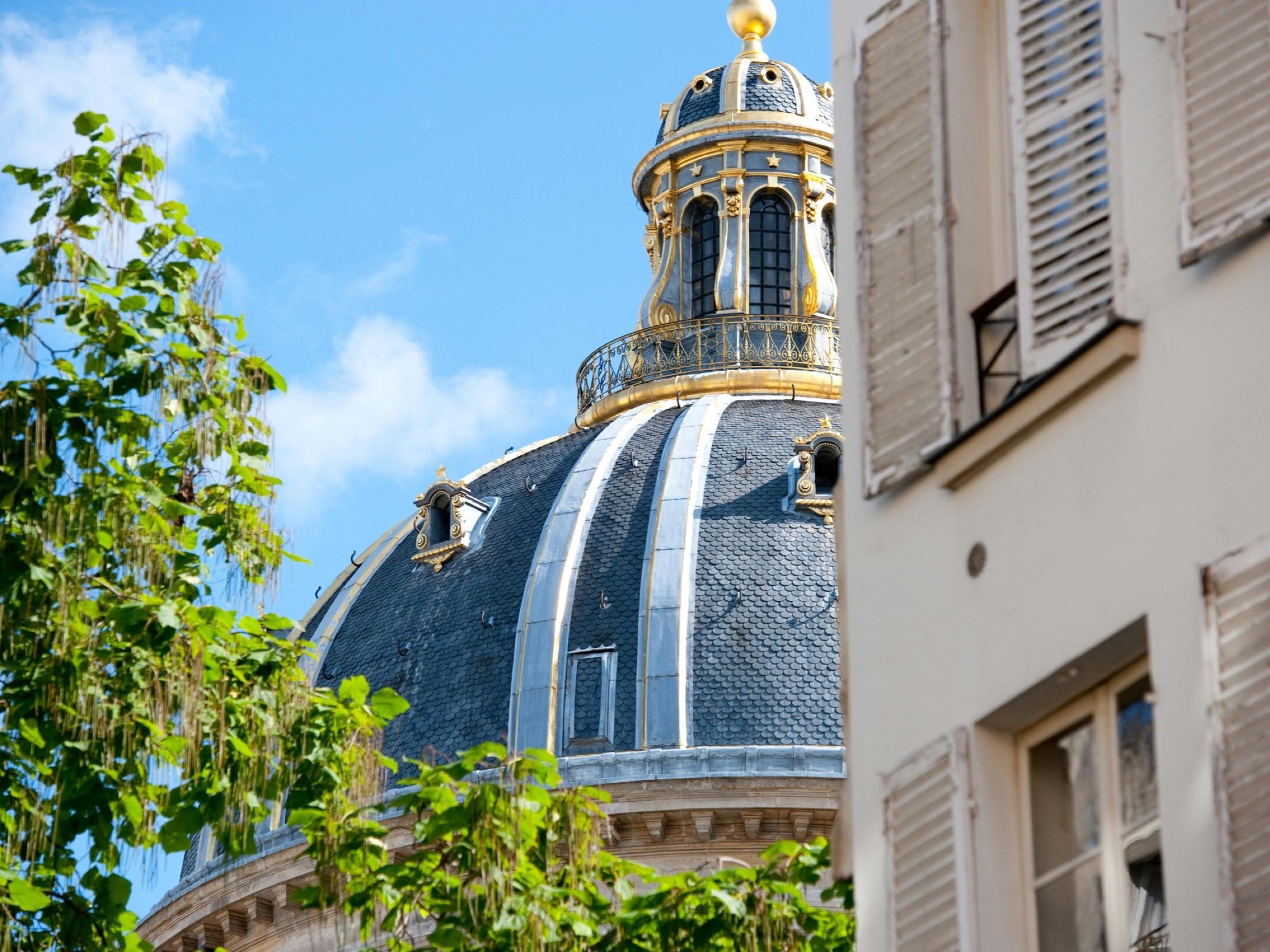 A Left Bank slate dome, its gilded lantern against a blue sky, seen past shuttered windows and a tree in leaf