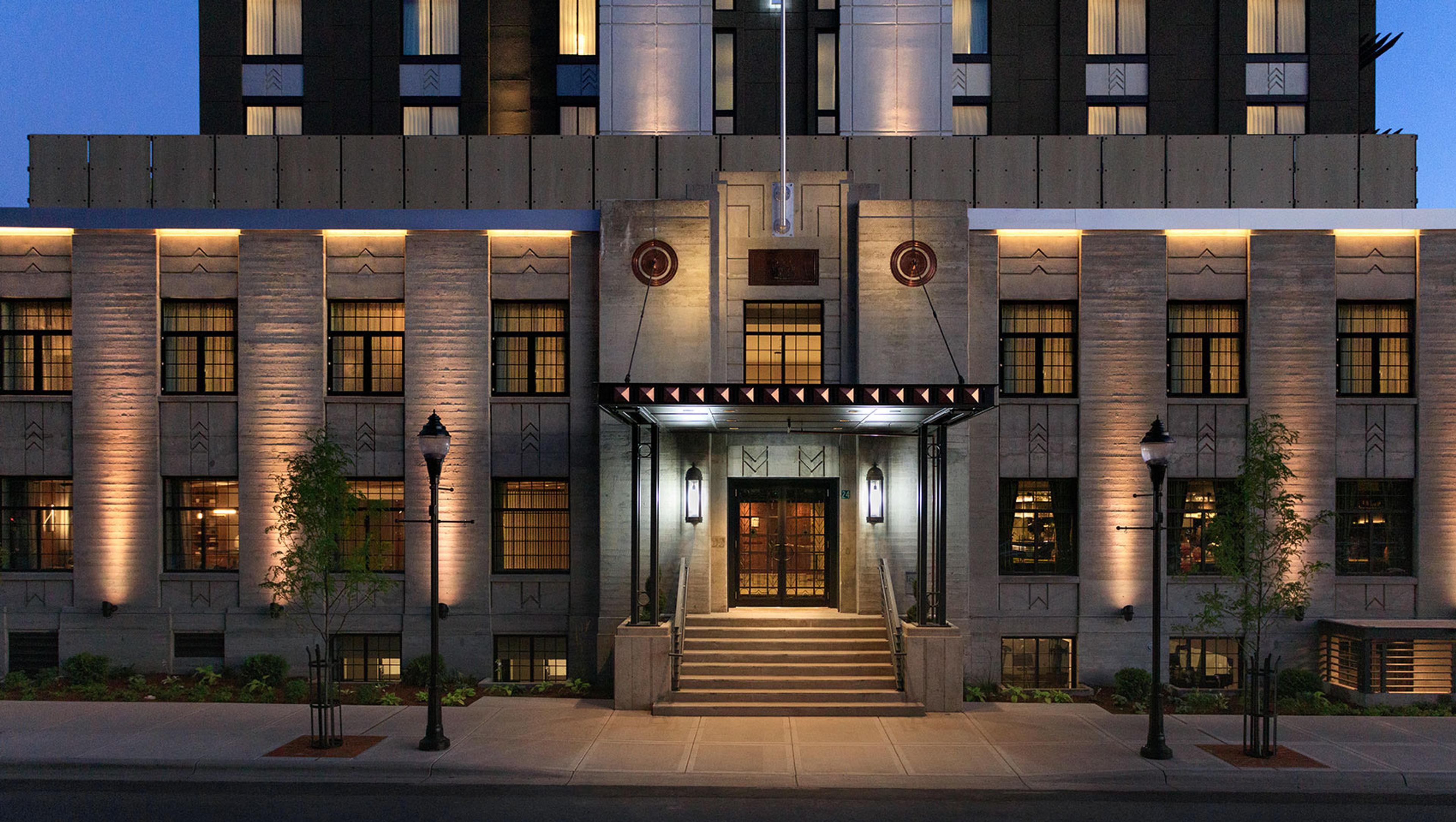 The hotel's stone facade lit up at dusk, with a covered entrance, a short flight of steps and two rows of tall windows glowing from inside.
