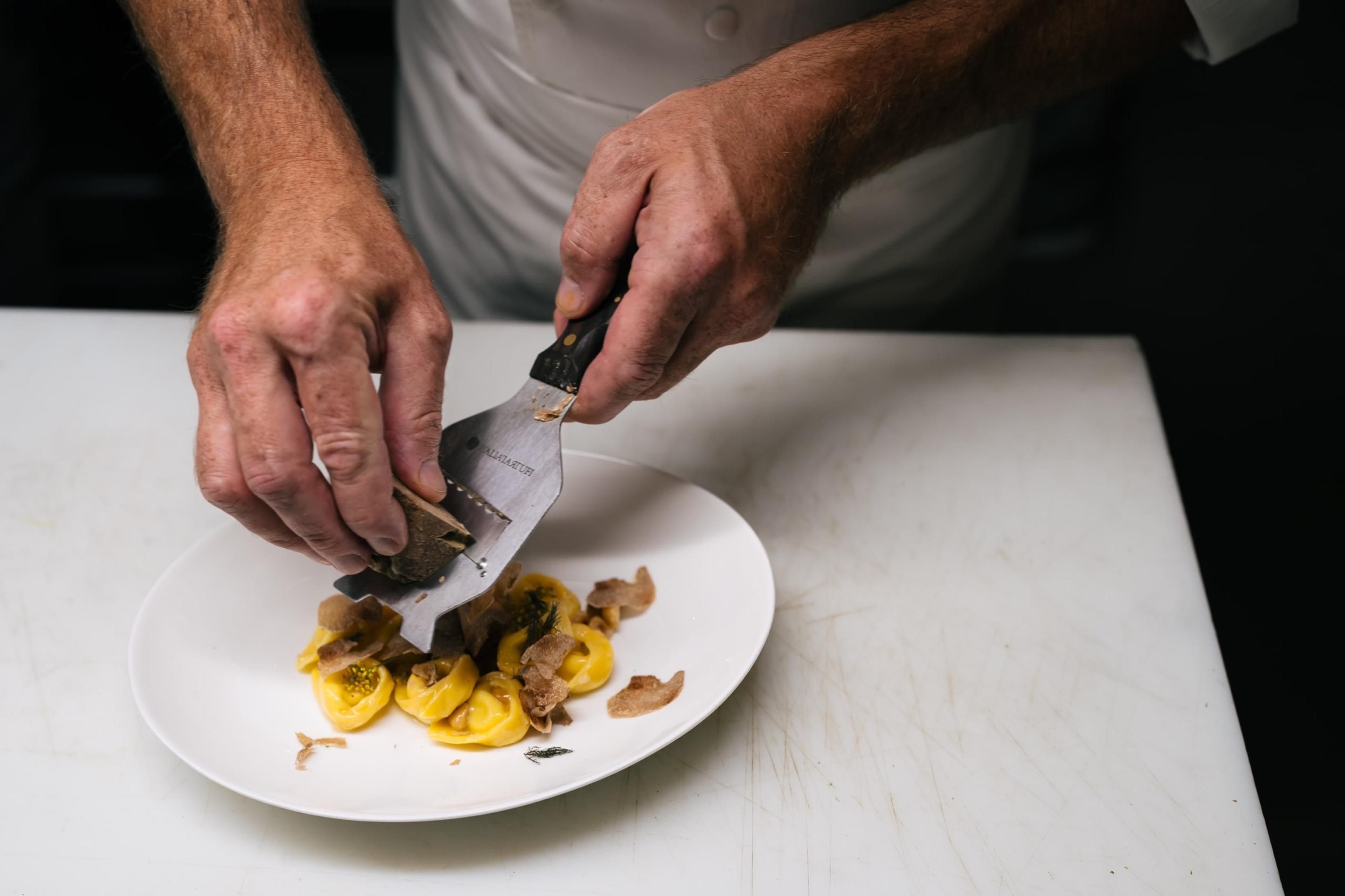 Two hands grate truffle over small yellow filled pasta on a white plate, on a white counter.