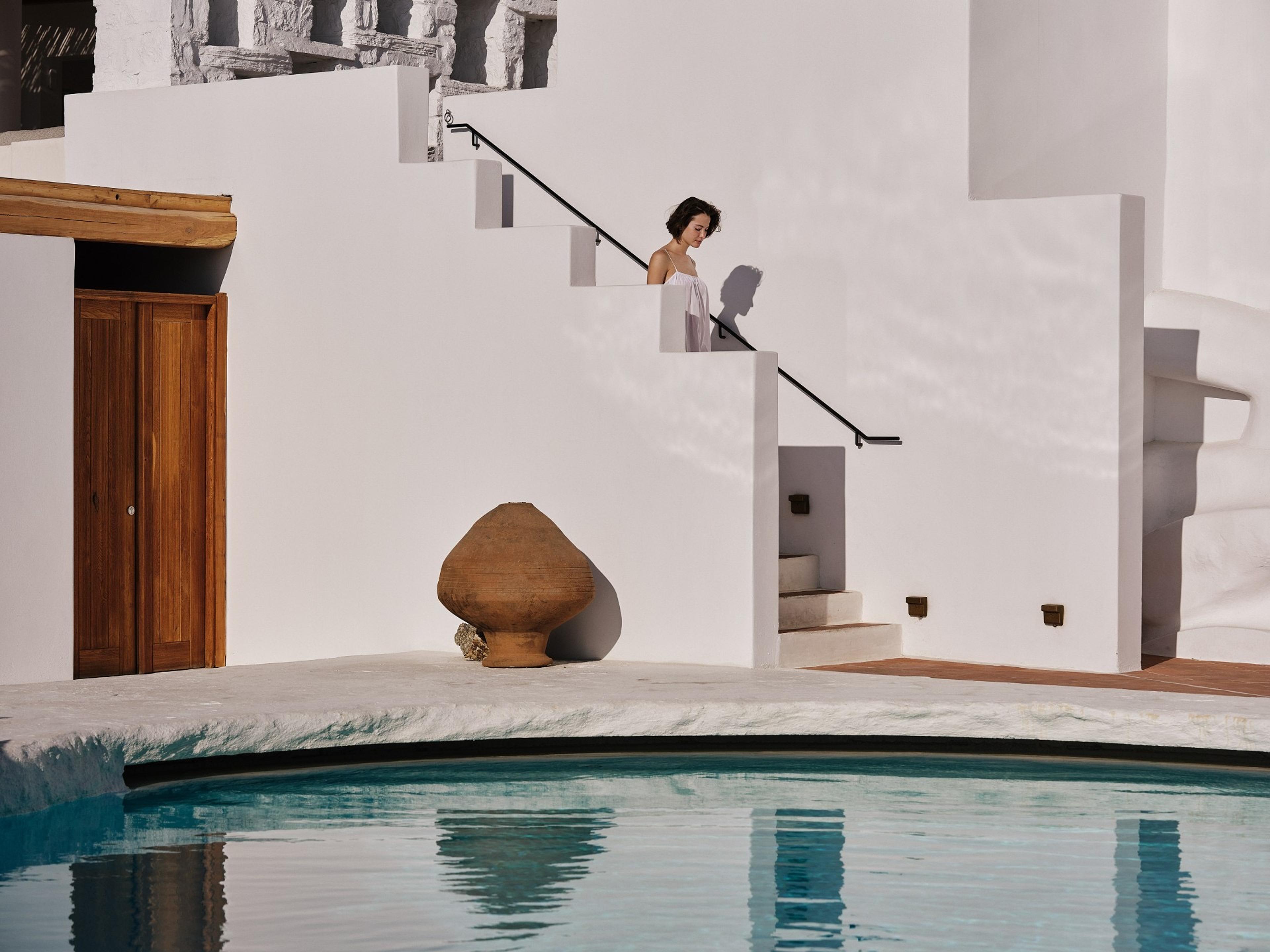 A woman in white walks down white stone steps past a large terracotta jar, reflected in a pool in front of her.