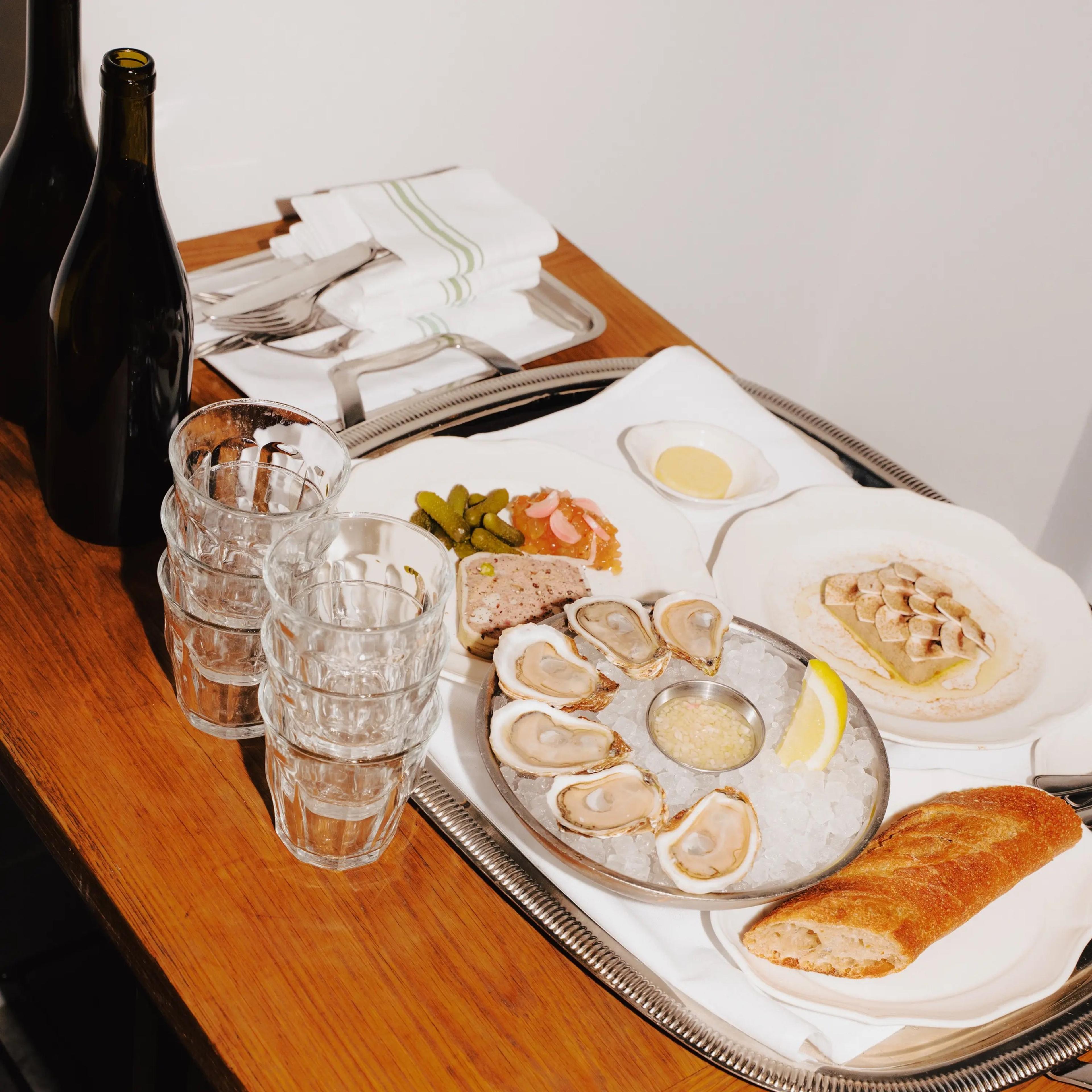 A room service tray on a wooden table with a dozen oysters on ice, pate, pickles, a baguette, and glasses of wine.